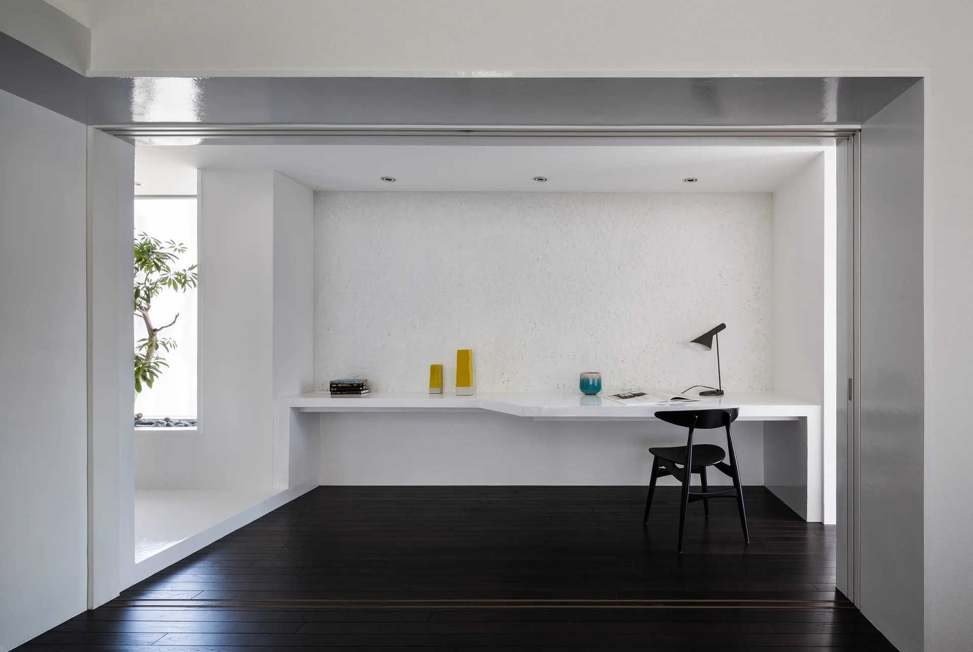 A minimalist home office features a long white built-in desk with a black wooden chair, a desk lamp, and dark wood plank floors.