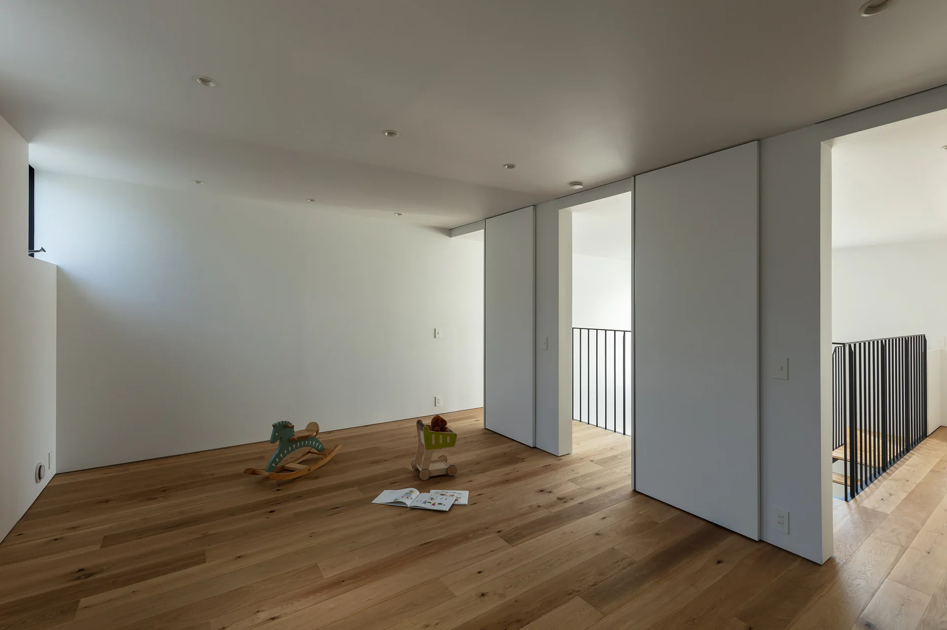 A minimalist playroom features light wood floors, white walls, sliding partition doors, and a wooden rocking horse.