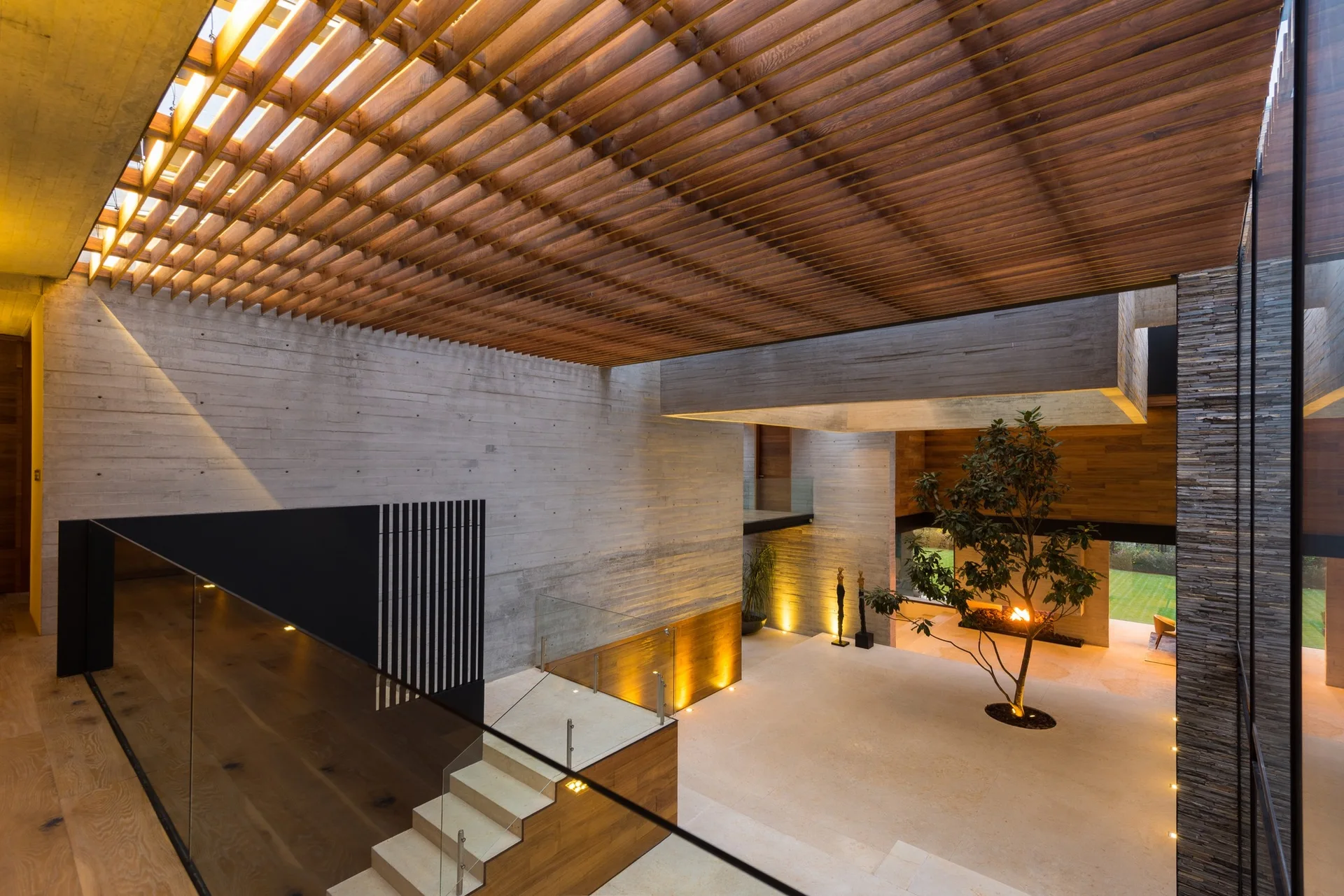 A modern spacious atrium featuring board-marked concrete walls, a wooden slatted ceiling overhead, a floating staircase, and a central indoor tree.