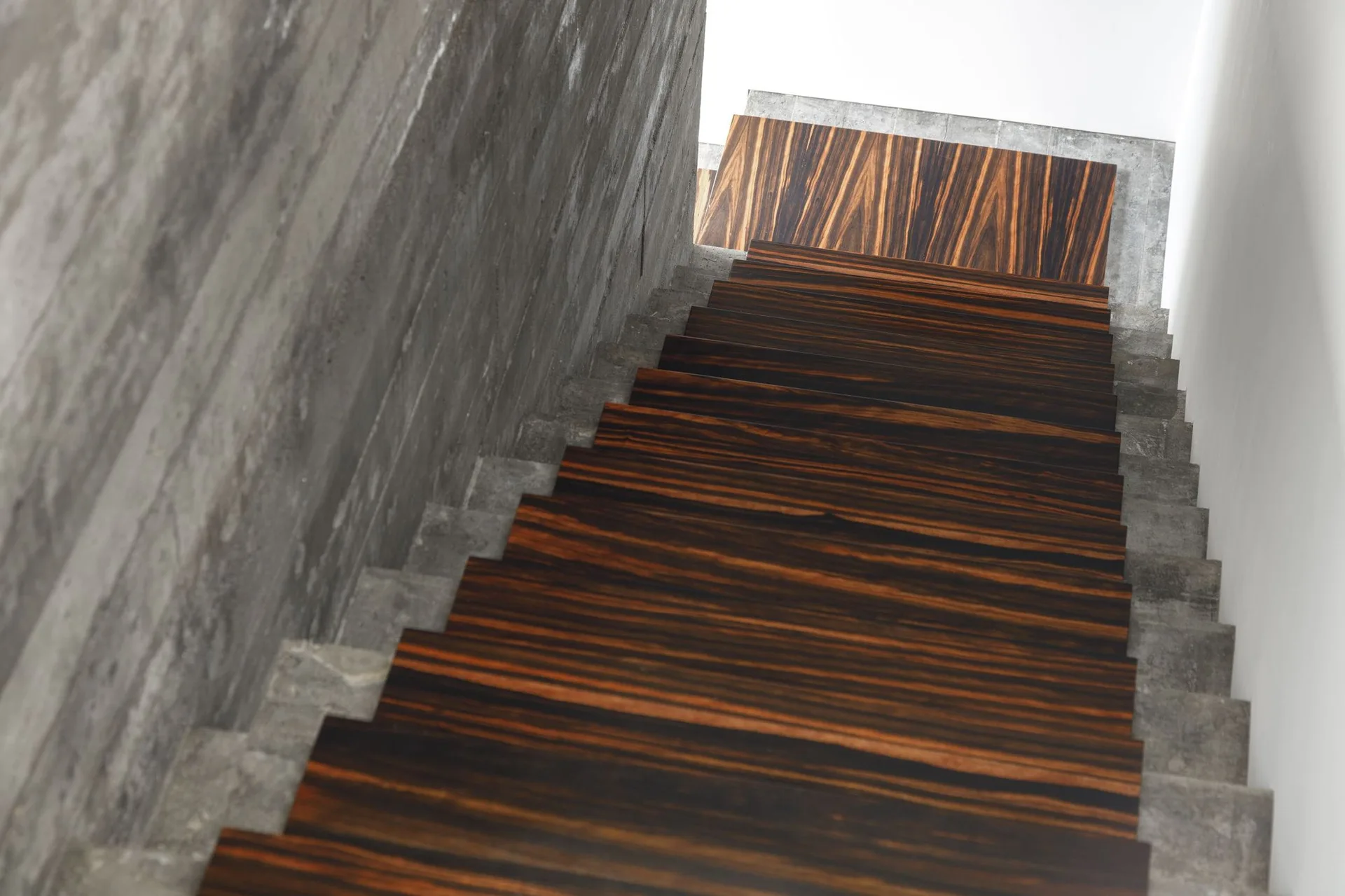 A high-angle view looking down a concrete staircase featuring rich dark brown wooden treads with a strong horizontal grain.