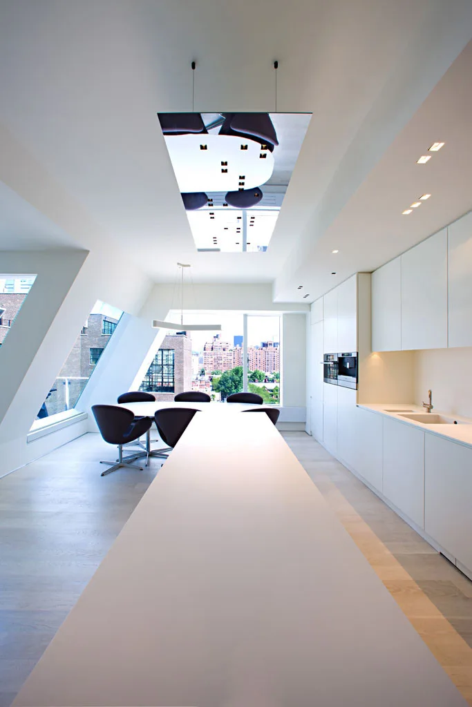 A minimalist white kitchen with a long central island, flush cabinetry, a dining table with dark chairs, and a slanted glass window wall.