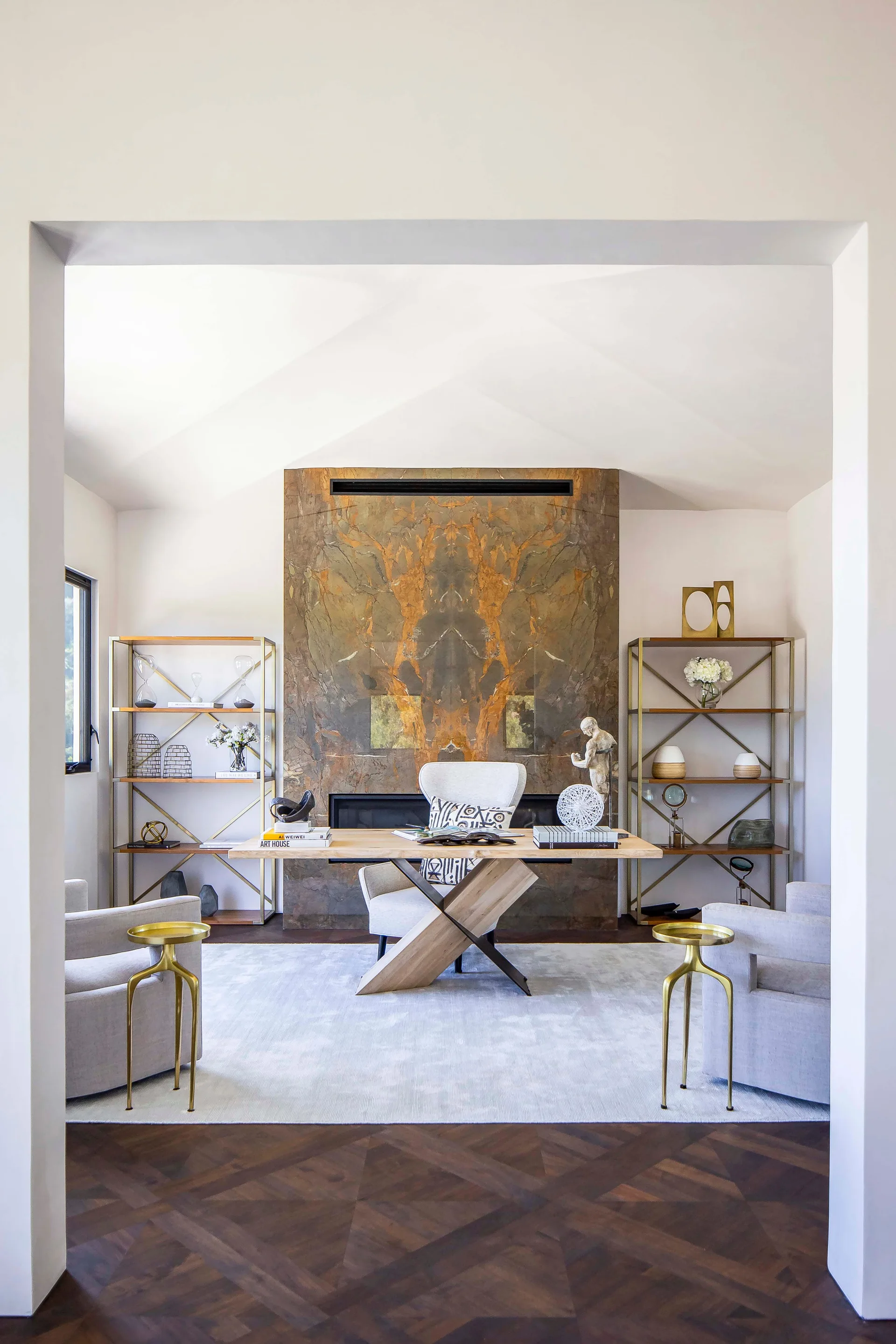 A symmetric home office featuring a central wooden desk in front of a tall stone fireplace, flanked by two brass open bookshelves.