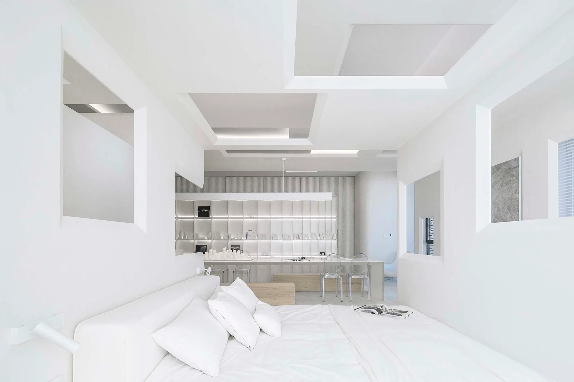A minimalist all-white bedroom features geometric ceiling cutouts, a platform bed, and backlit built-in shelving in the background.