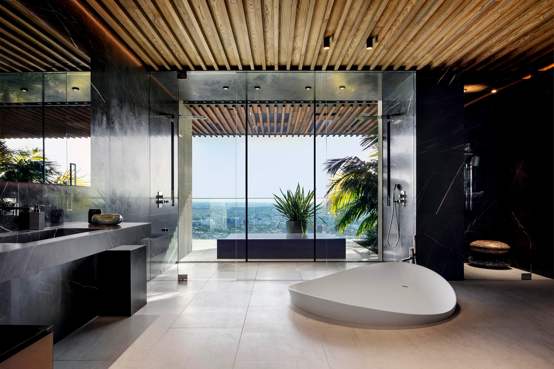 A modern luxury bathroom features dark stone walls, a slatted wood ceiling, and a low-profile organic white tub facing a glass wall with city views.