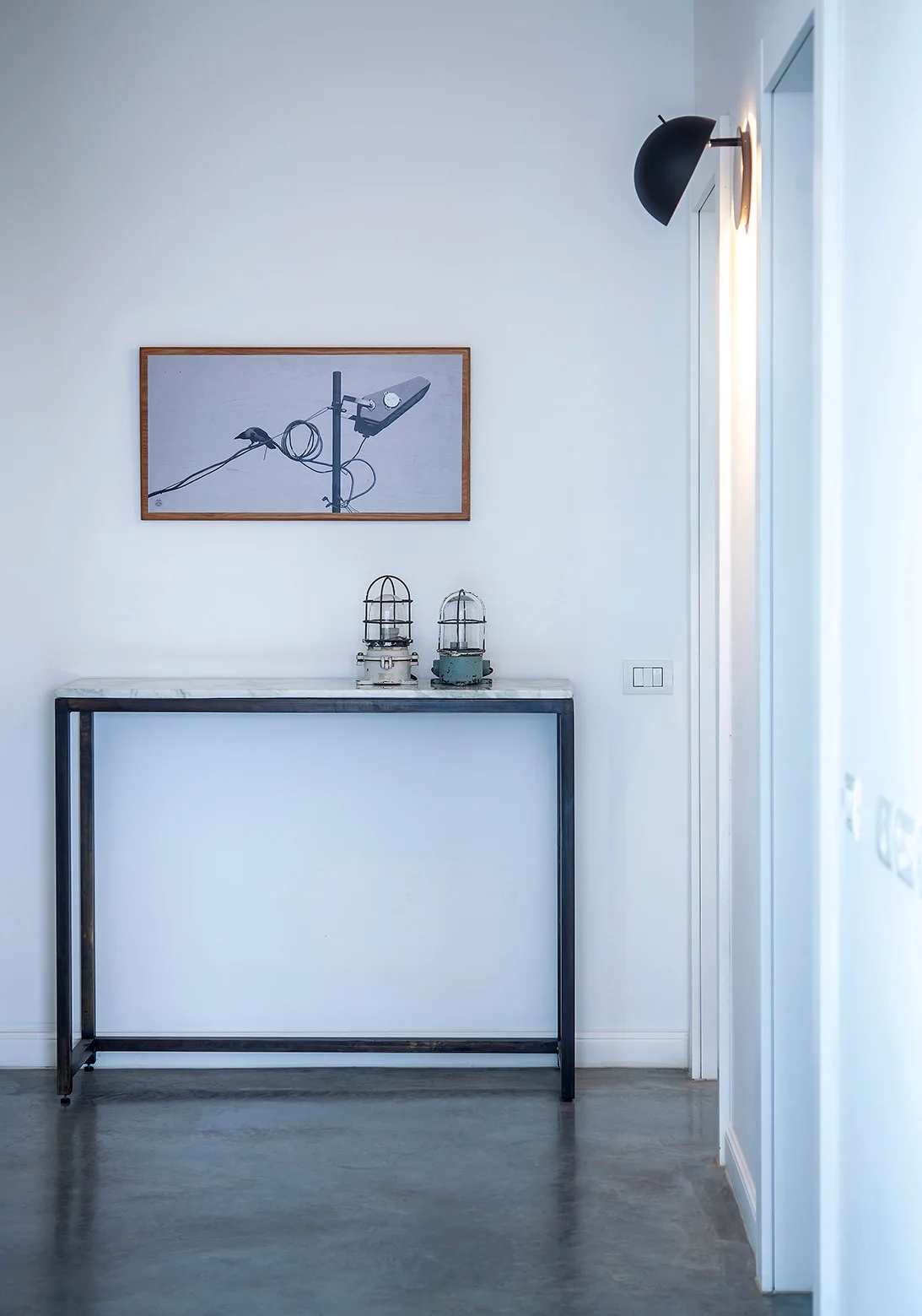 A slim console table with a stone top holds two caged metal lamps against a white wall next to a black sconce lamp.