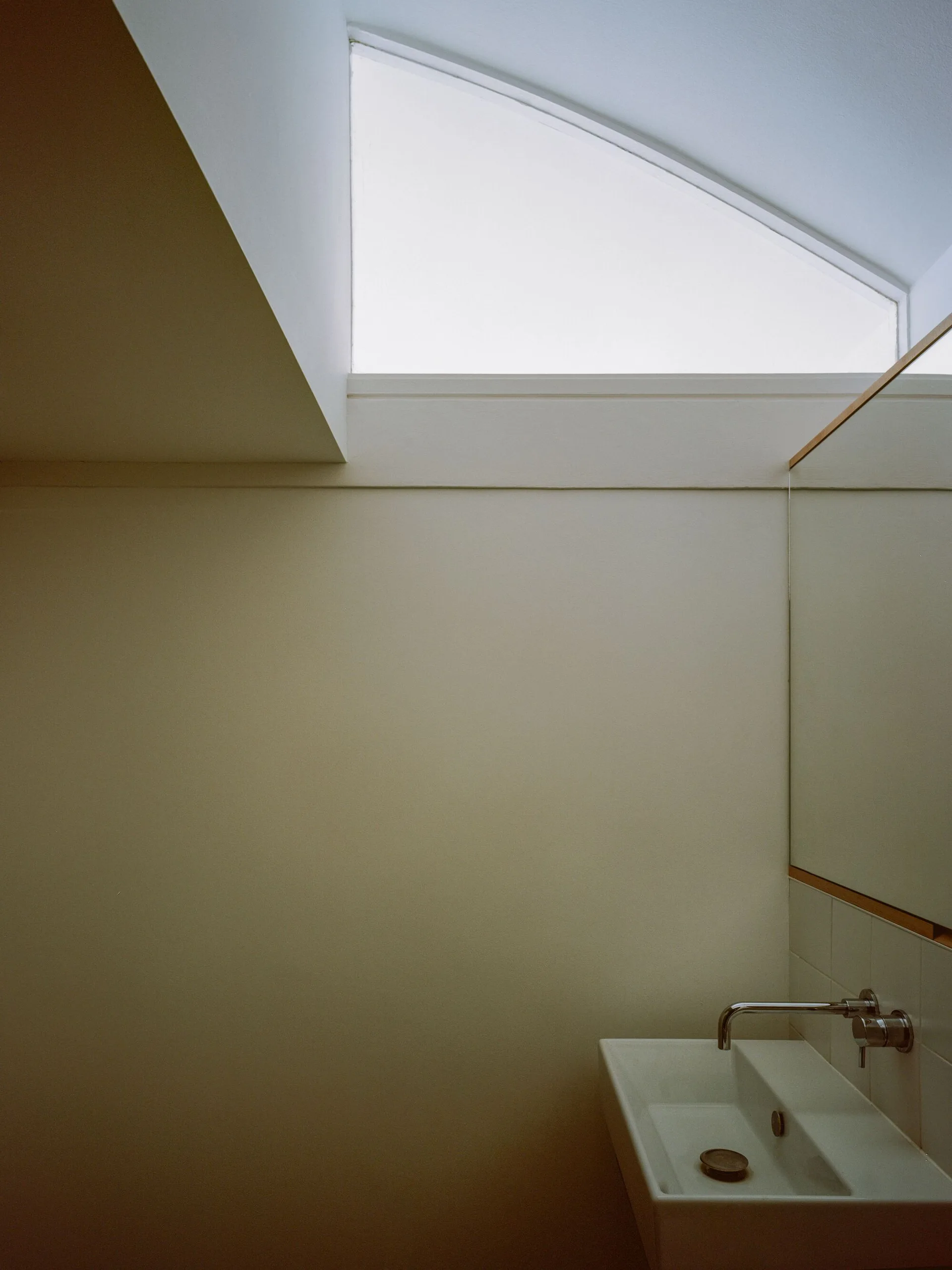A minimalist bathroom featuring a wall-mounted ceramic sink with a metal faucet, a wood-trimmed mirror, and an angled high window.