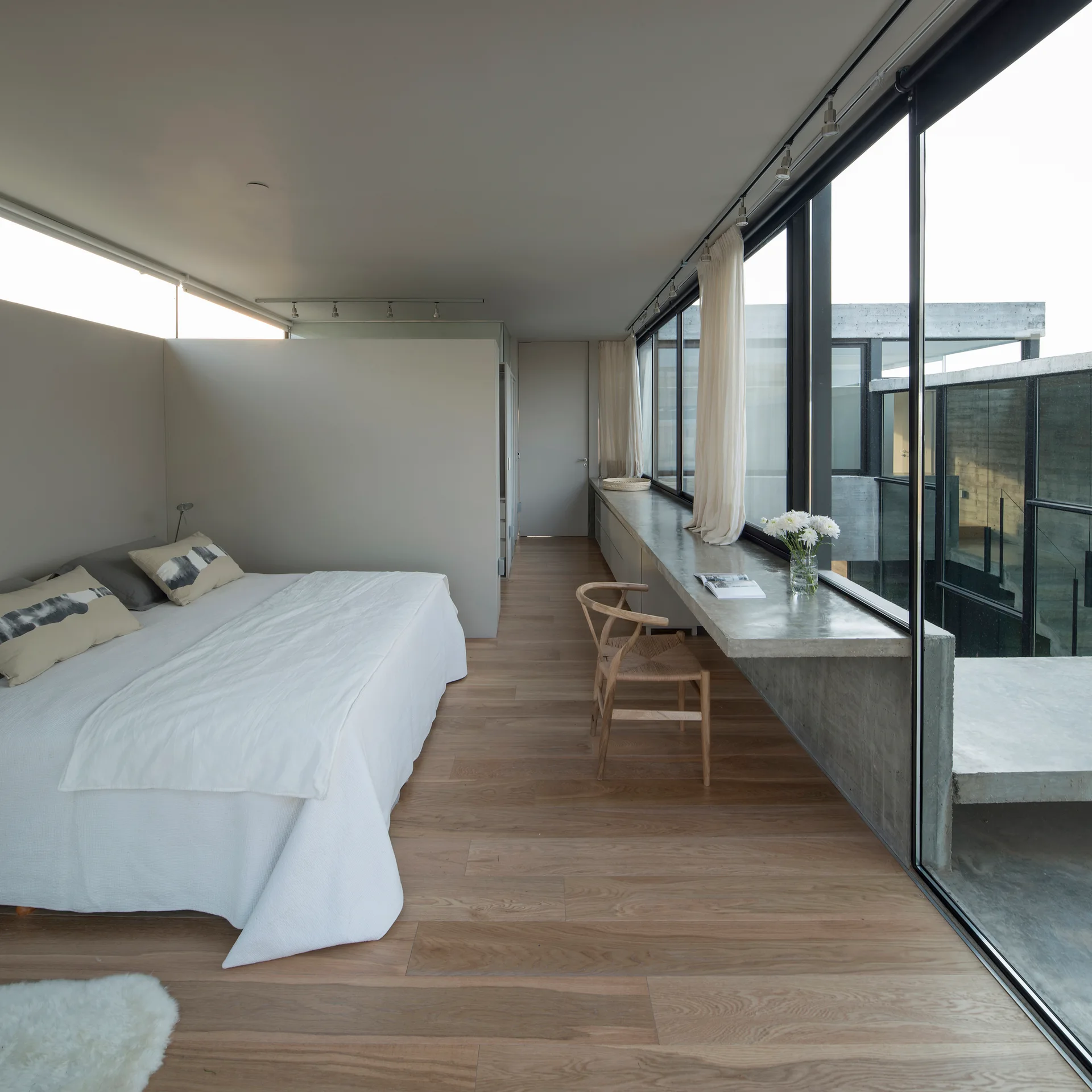 A minimalist bedroom featuring a bed with white linens and a built-in concrete desk along a large wall of windows.
