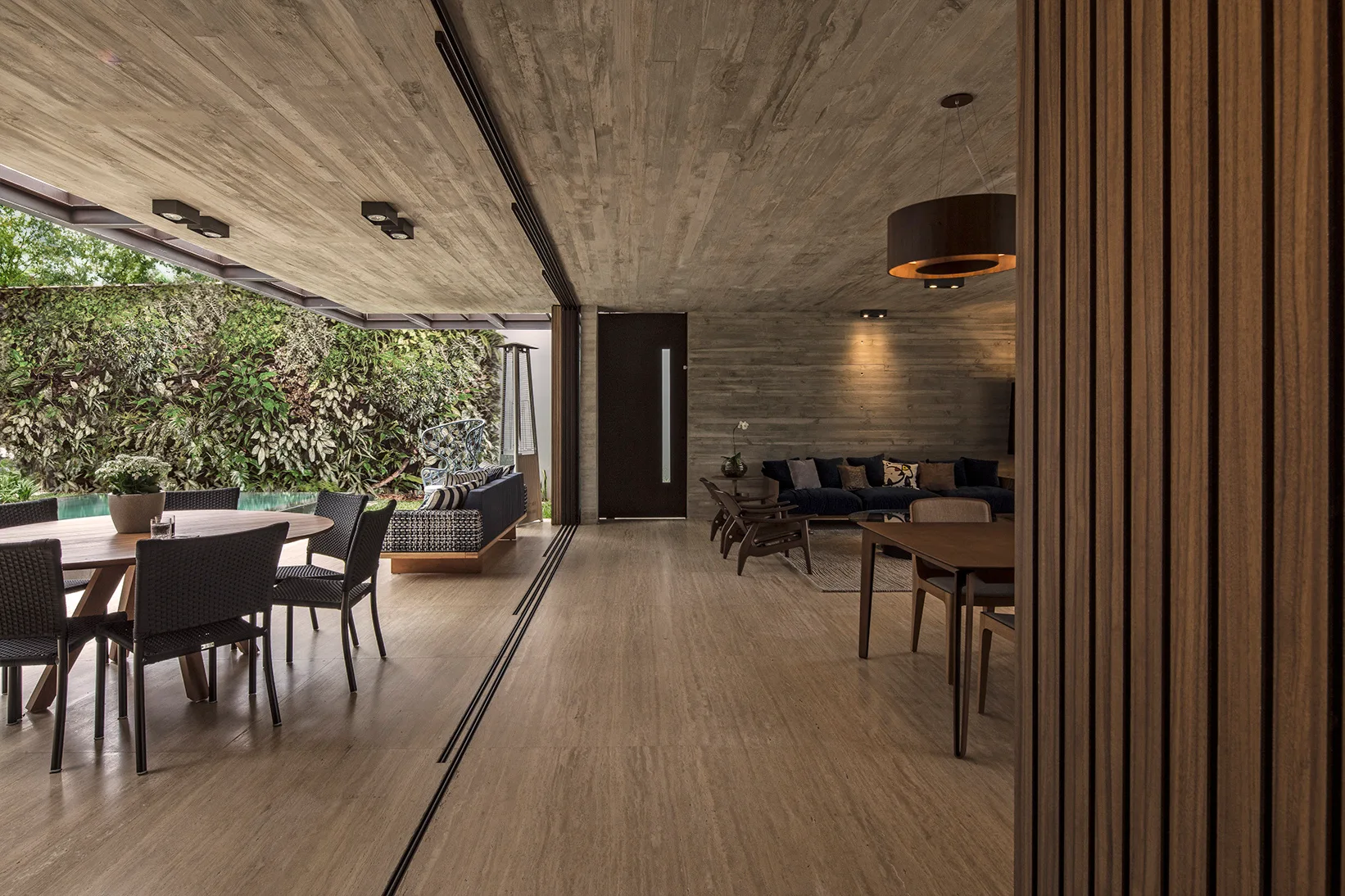 A modern open-plan patio and living room with board-formed concrete ceilings, a round wood dining table, and large sliding doors opening to a lush garden.