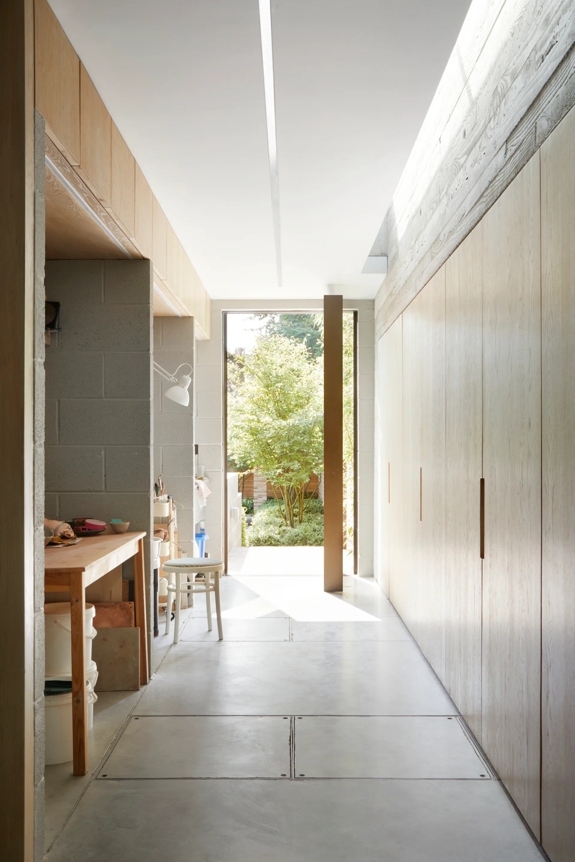 A minimalist hallway features light wood built-in cabinetry, textured concrete block walls, and a glass door opening to an outdoor garden.
