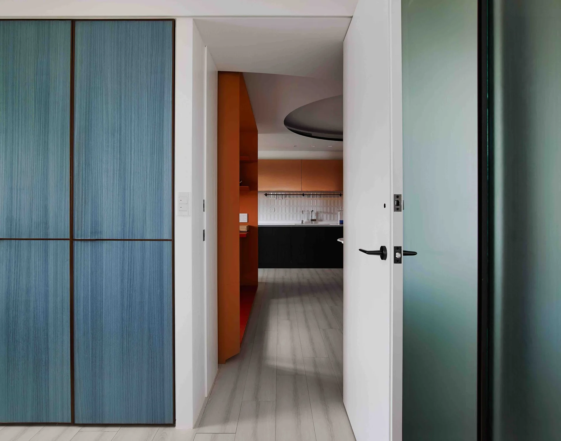 A white doorway opens to a hallway with blue textured wood panels leading toward a kitchen with orange cabinets.