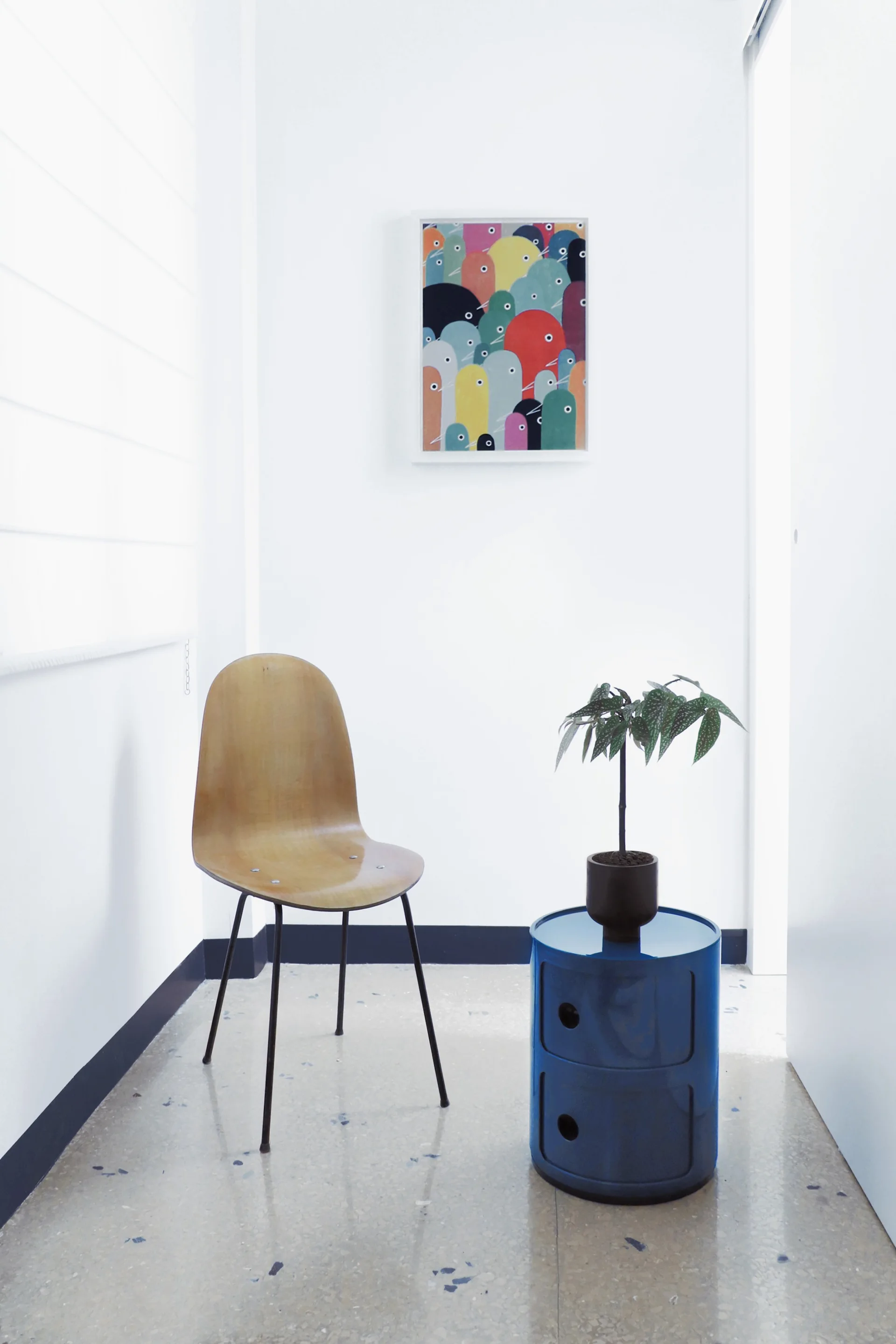 A molded plywood chair sits next to a glossy blue cylindrical storage table with a small plant in a minimalist white-walled corner.
