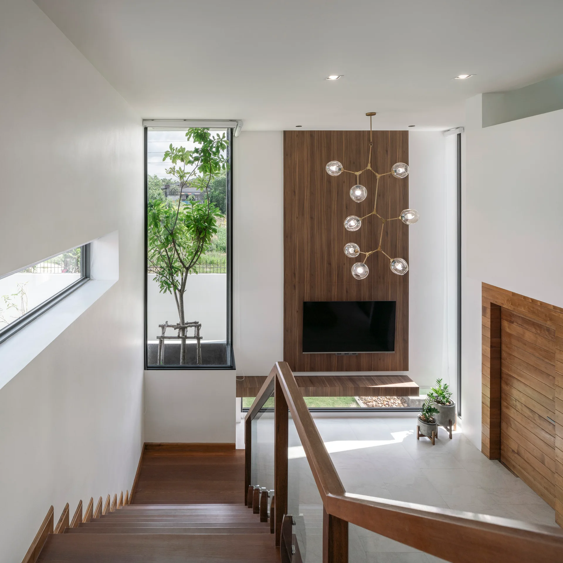 A wooden staircase with a glass railing overlooks a white living space with a wood-paneled TV feature wall, tall window, and branching globe chandelier.