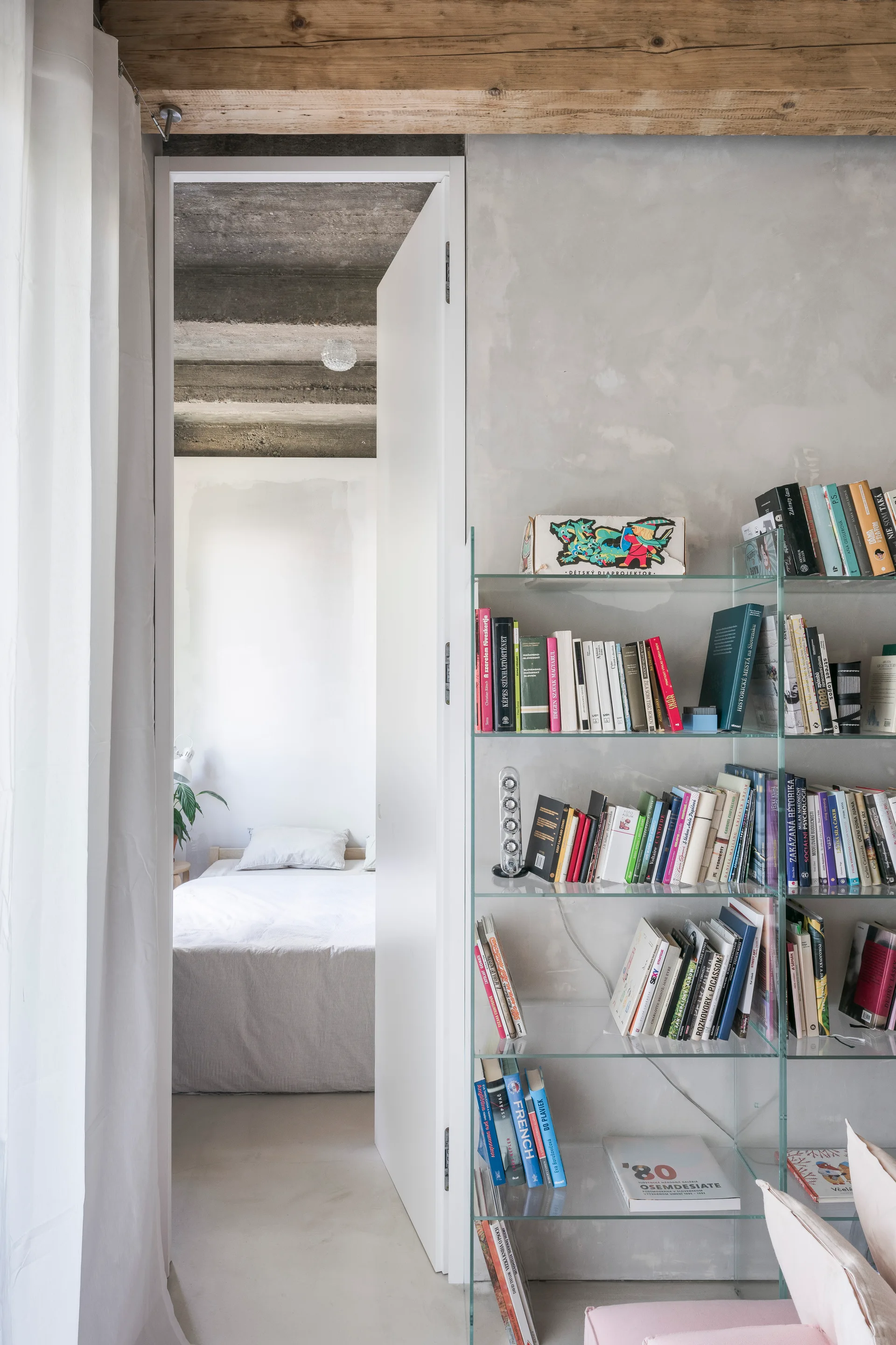 An open white doorway reveals a minimalist bedroom framed by a clear glass bookshelf stocked with books against a gray textured plaster wall.