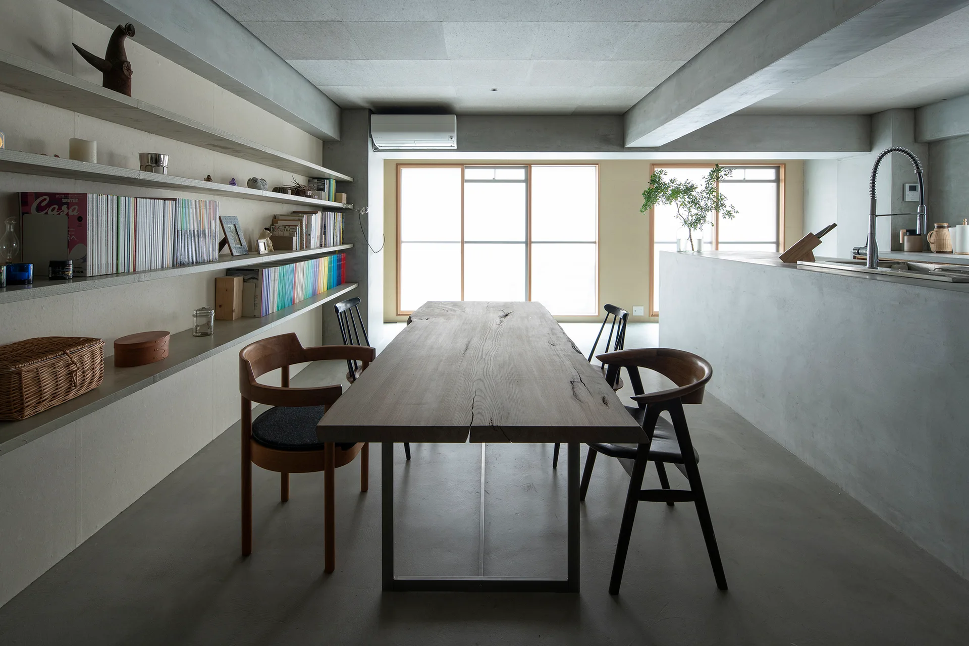 A minimalist dining room featuring a live-edge wooden table, concrete floor, concrete island, and long open wall shelves with books.