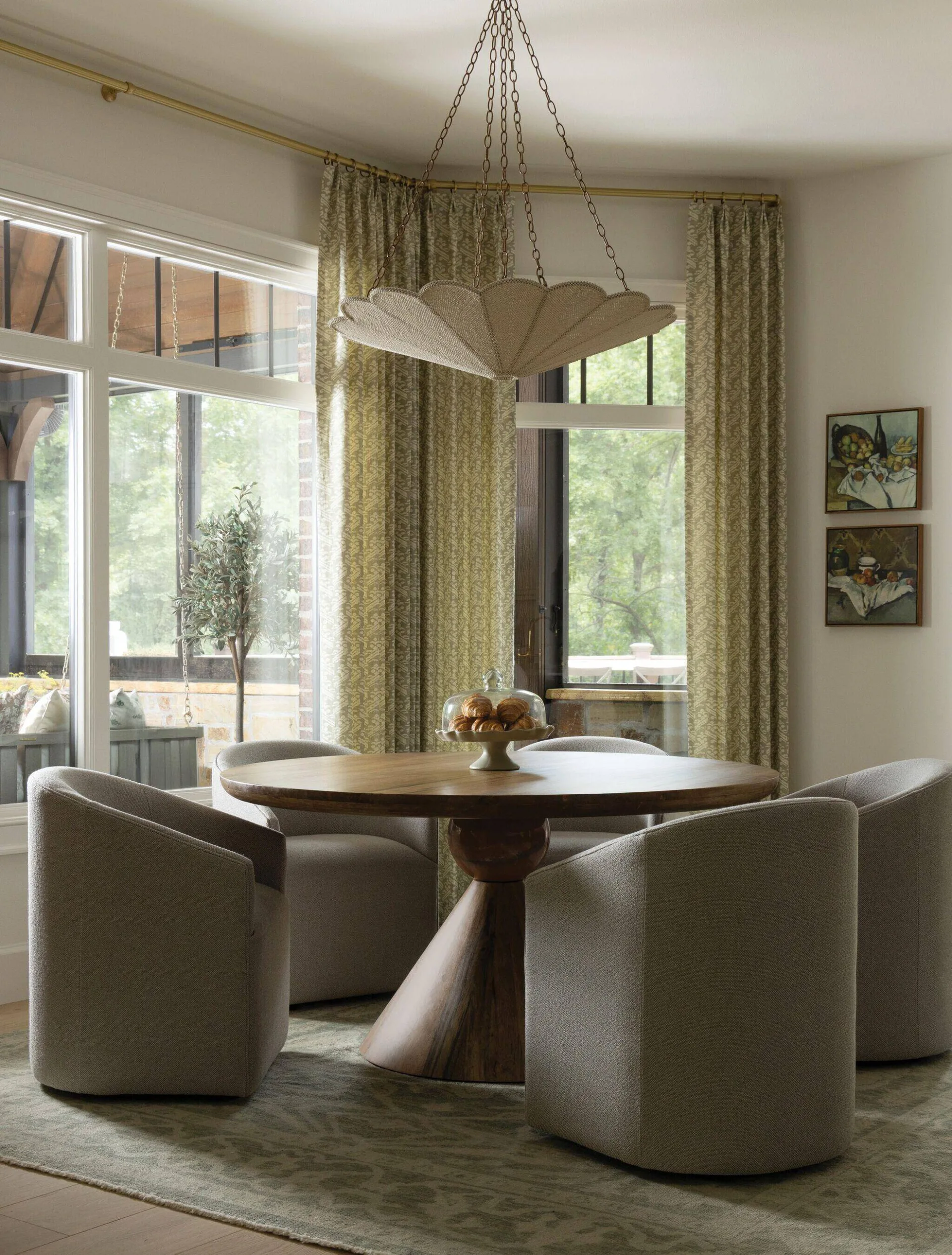 A round wooden pedestal dining table surrounded by four beige upholstered barrel chairs stands on a patterned rug near a window with sage green curtains.