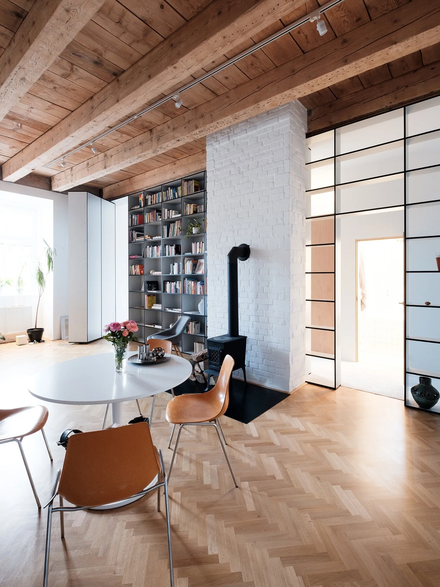 A modern dining space with a round white table, wooden chairs, a white brick fireplace column, and exposed wooden ceiling beams.