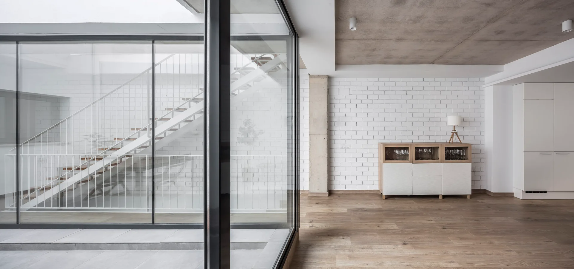 A minimalist living area features an open-riser staircase behind a glass partition, an exposed concrete ceiling, and a white brick wall.