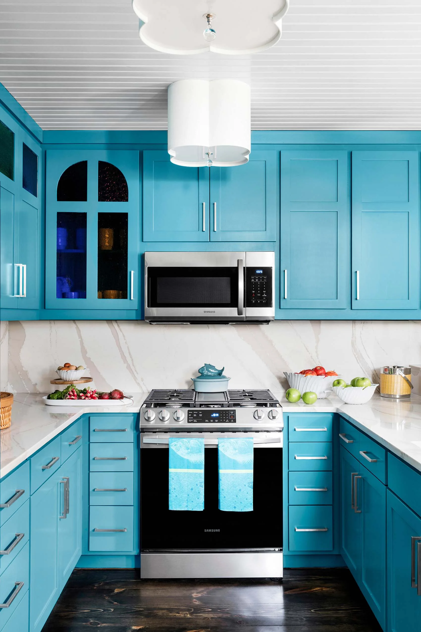 A modern kitchen features vibrant turquoise cabinets, a veined white stone backsplash, stainless steel appliances, and a white beadboard ceiling.