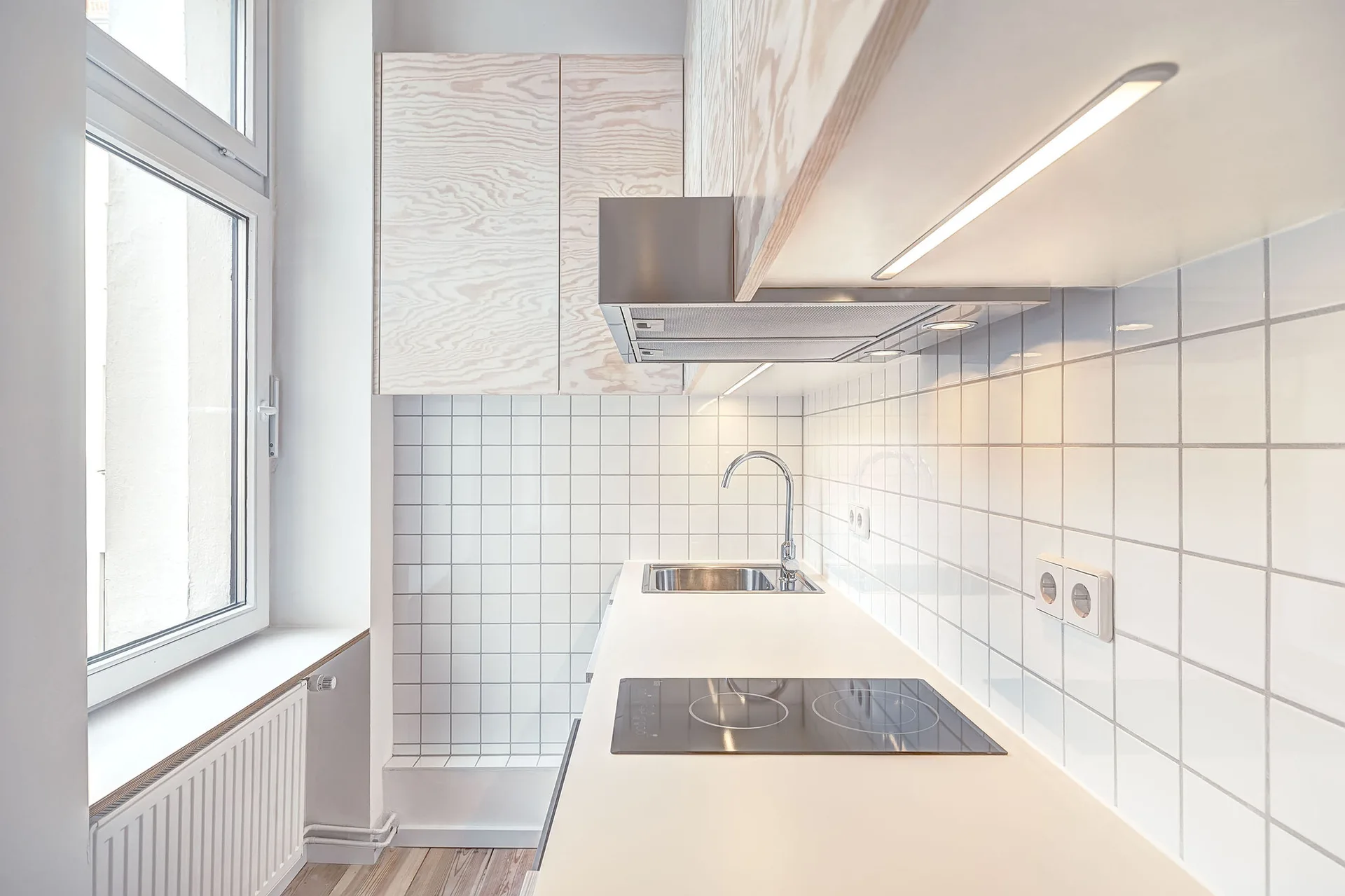 A minimalist kitchen featuring light wood grain cabinets, white square grid tile backsplash, and a white countertop with an integrated stovetop and sink.