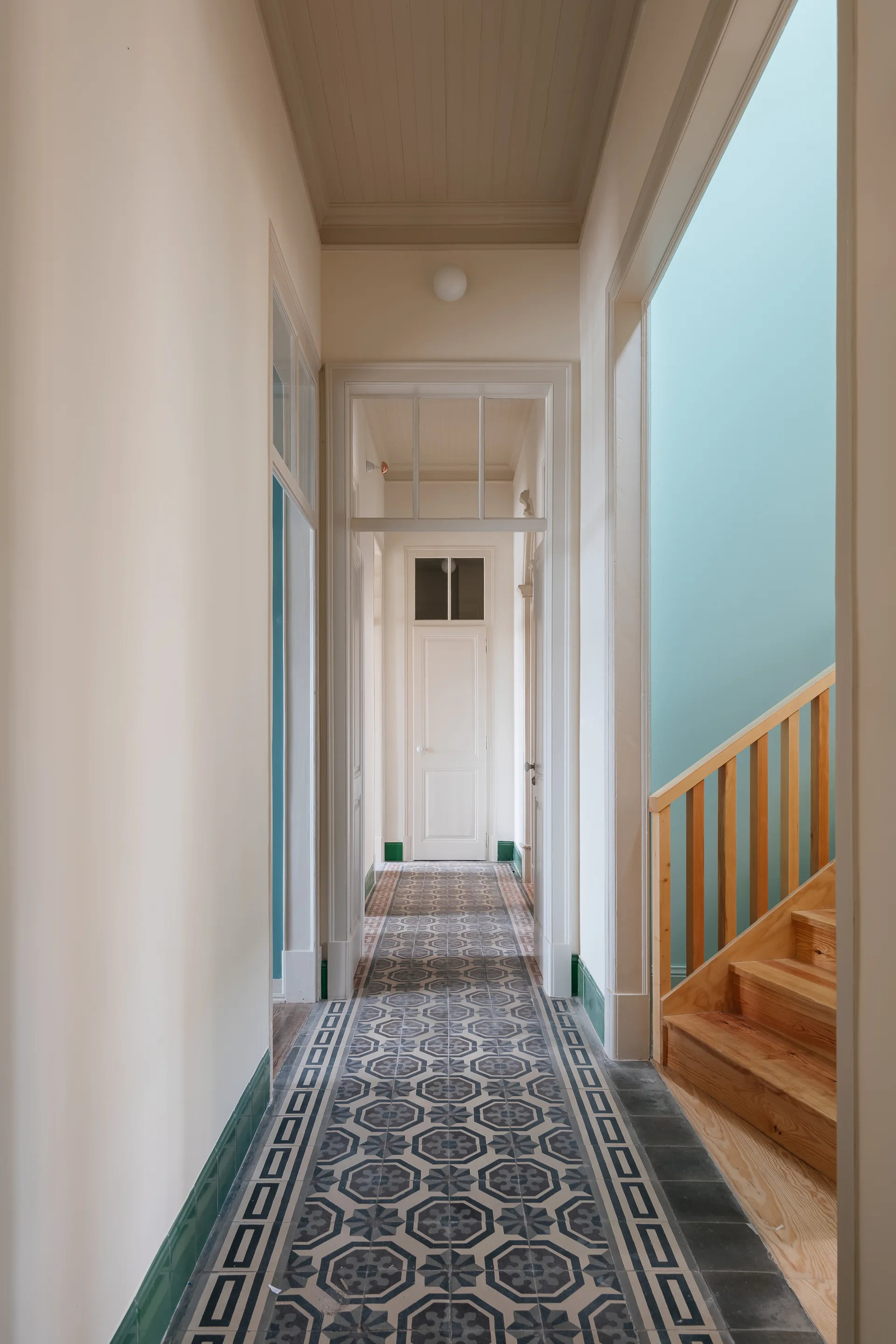 A long hallway featuring geometric patterned floor tiles, white walls with glossy green baseboard trim, and a wooden staircase on the right.