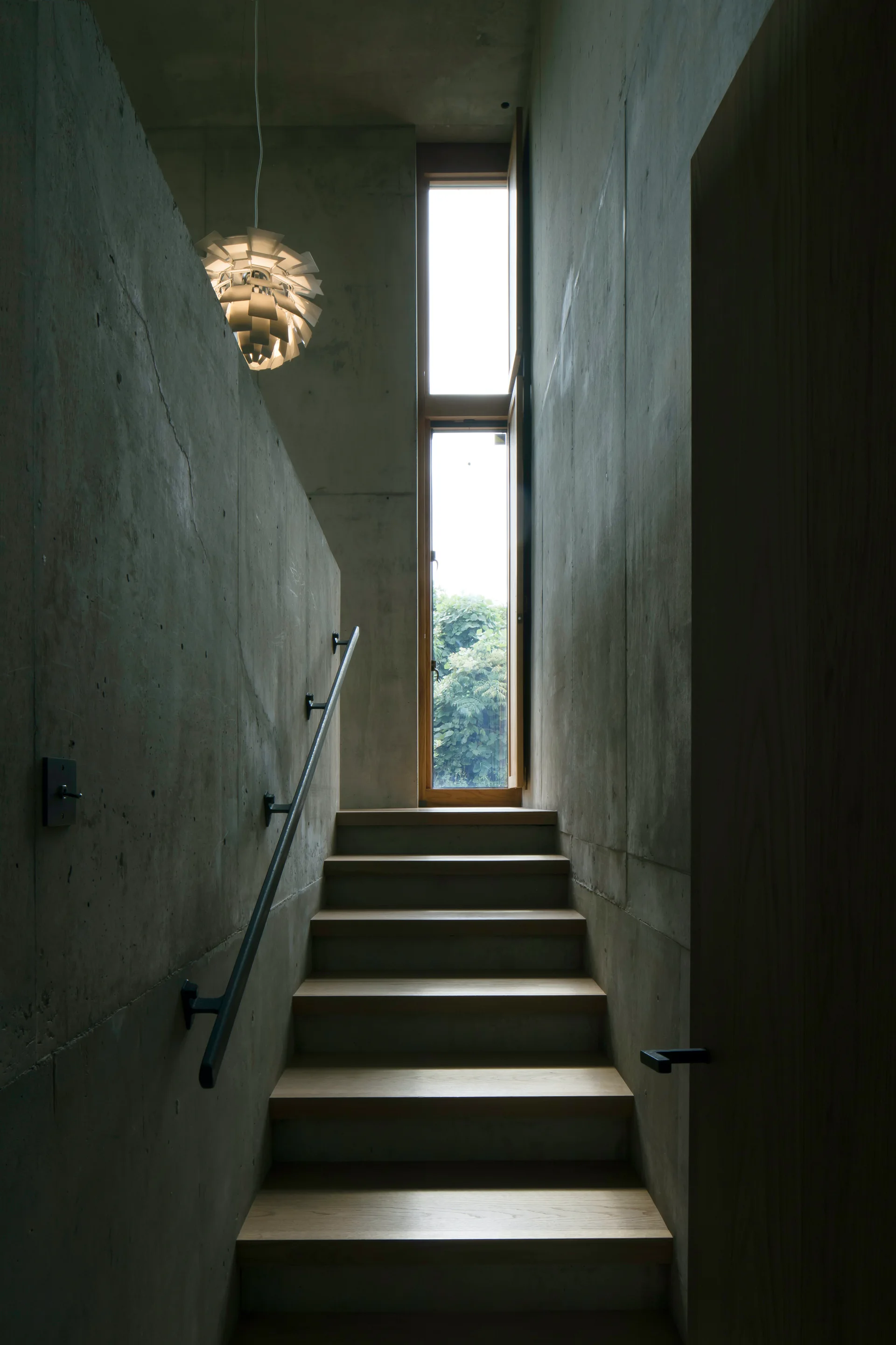 A modern staircase featuring wooden treads, a dark metal handrail, raw concrete walls, a tall narrow window, and a hanging white pendant light.