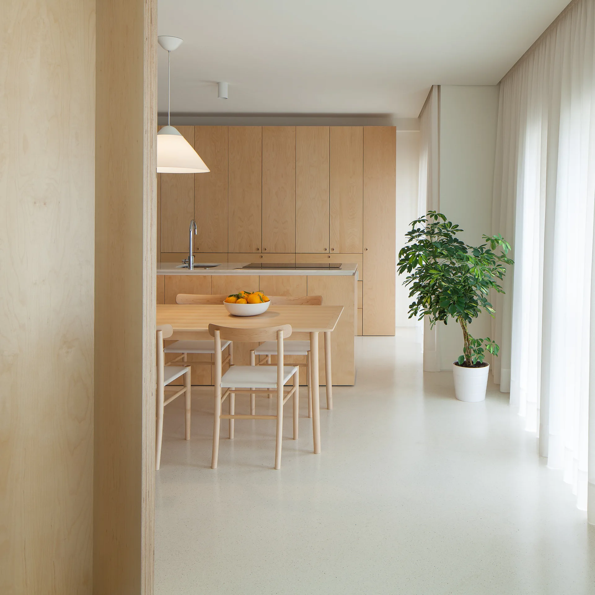A minimalist kitchen and dining area featuring light wood cabinetry, a matching dining table, sheer white curtains, and a potted plant.