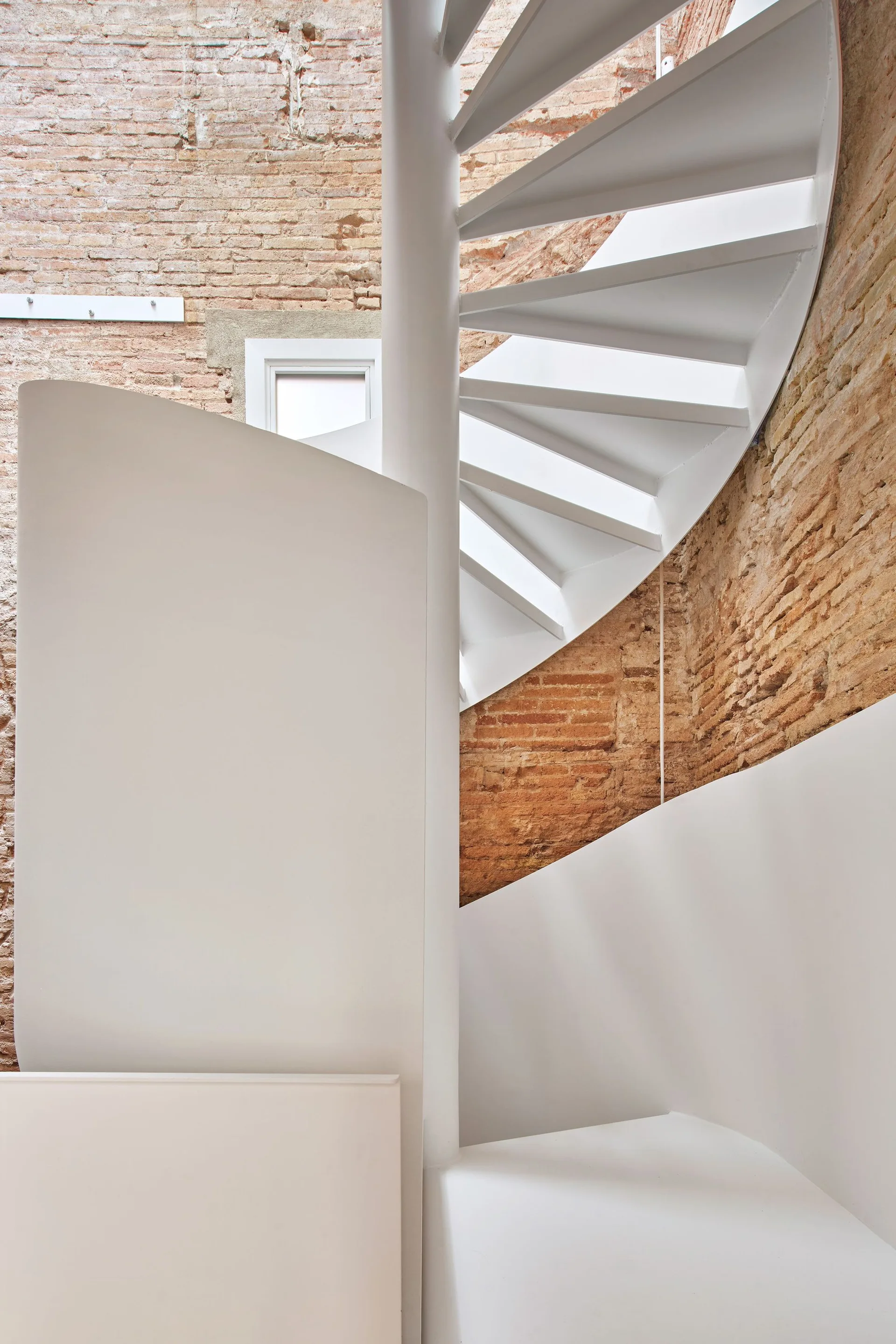 A minimalist white metal spiral staircase ascends next to a textured, exposed red brick wall with a small window visible behind.