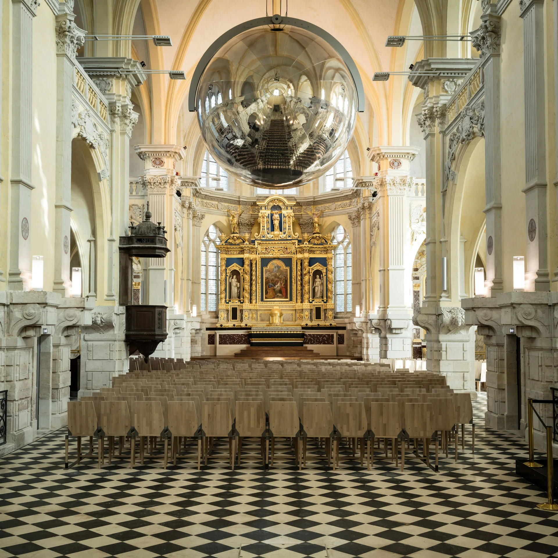 Rows of light wood chairs face an ornate gold altar beneath a large reflective silver sphere suspended from a vaulted ceiling.