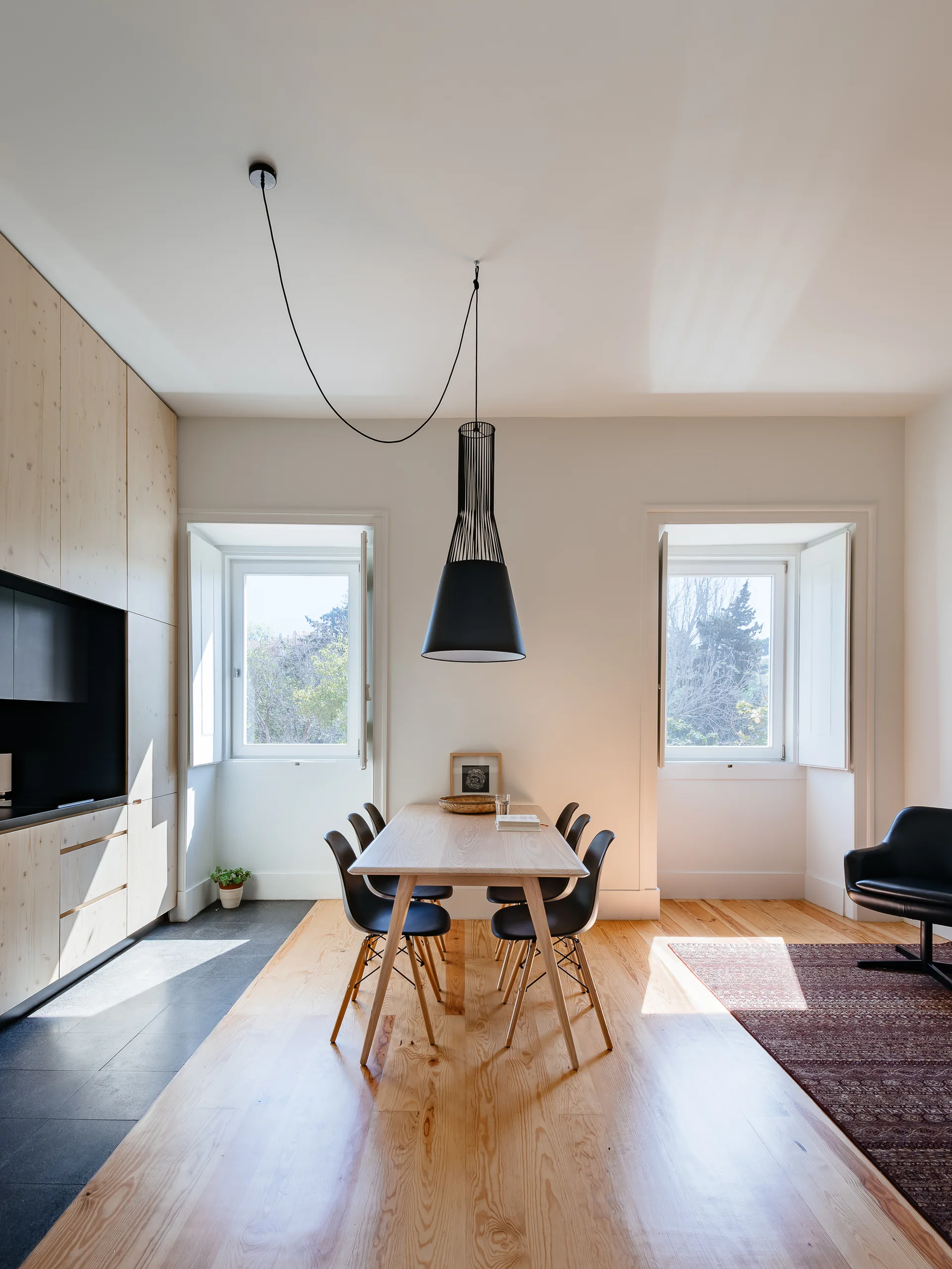 A bright Scandinavian dining room features a light wood table, black chairs, a black pendant light, and a wall of matching light wood cabinetry.
