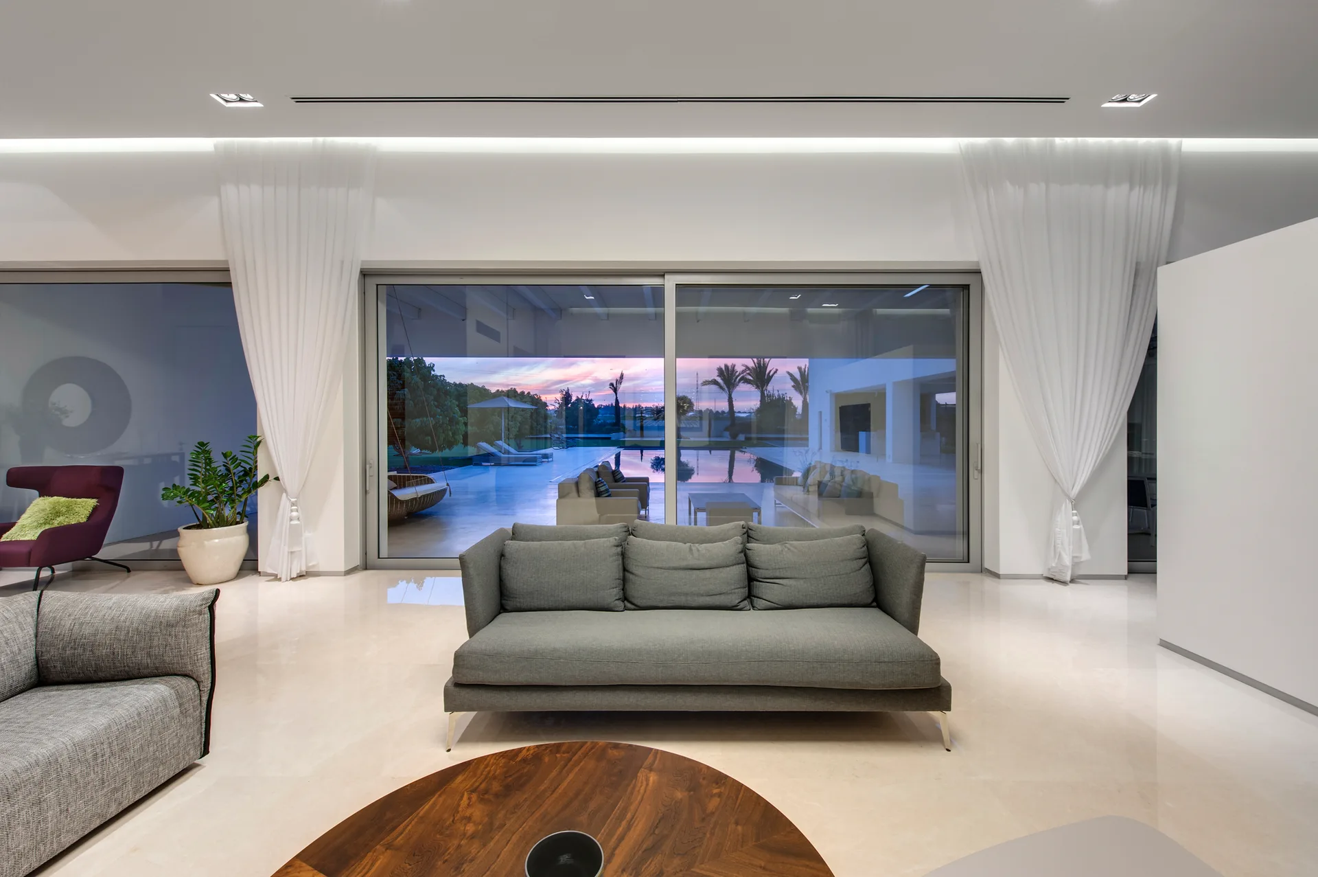 A minimalist living room features a grey sofa, polished beige floors, and large sliding glass doors overlooking an outdoor pool at sunset.