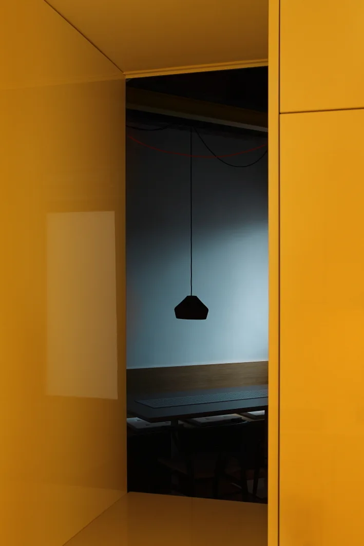 A view through a bright yellow glossy doorway looking into a dimly lit dining area with a dark wood table and a black pendant light.