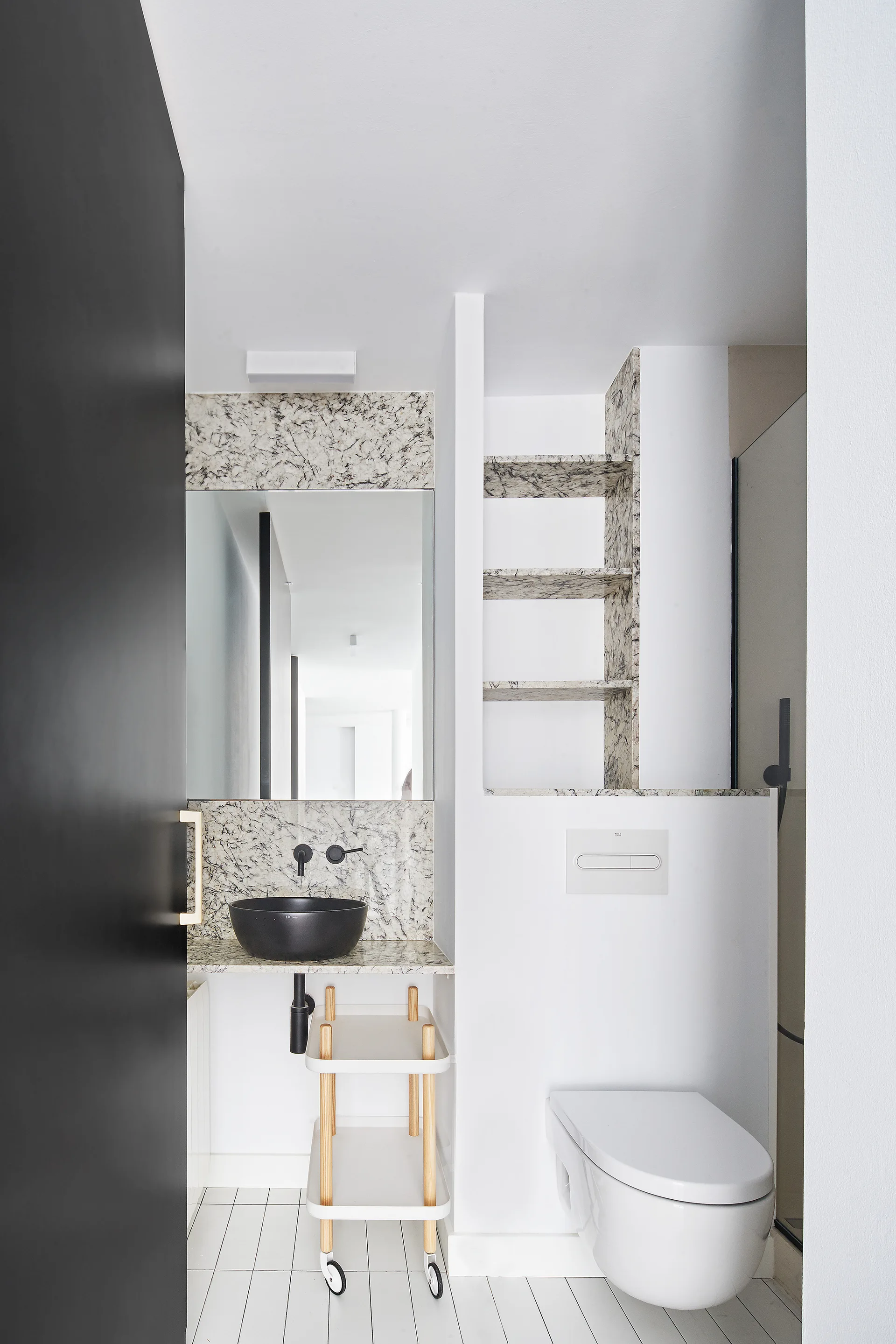 A minimalist bathroom featuring a black vessel sink, marble-veined backsplash, recessed stone shelves, a wall-mounted toilet, and a black door.