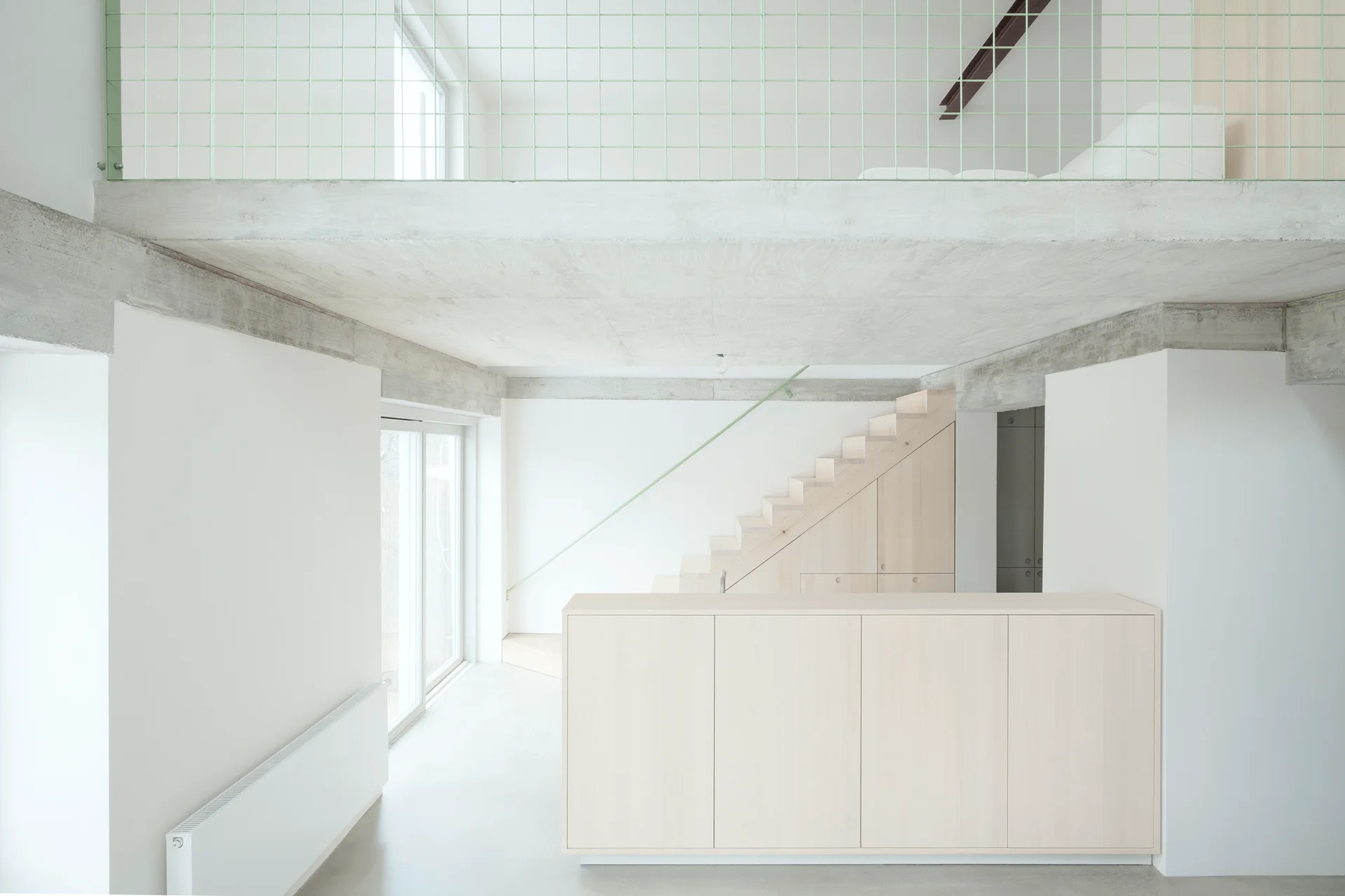 A minimalist loft space features an integrated light wood staircase, a matching foreground cabinet, a raw concrete ceiling, and a mint green wire grid railing.