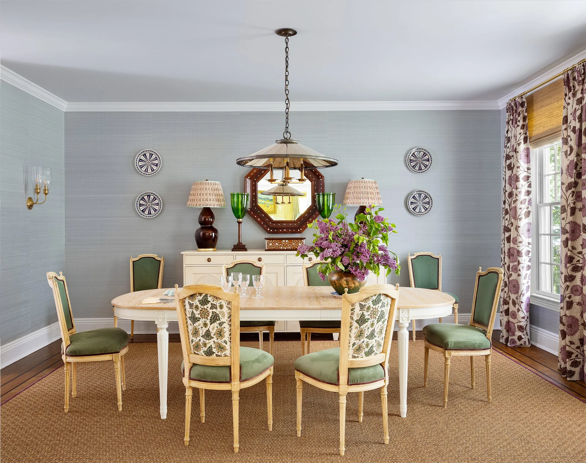 A formal dining room features a pale wood oval table surrounded by green chairs against light blue grasscloth wallpaper.