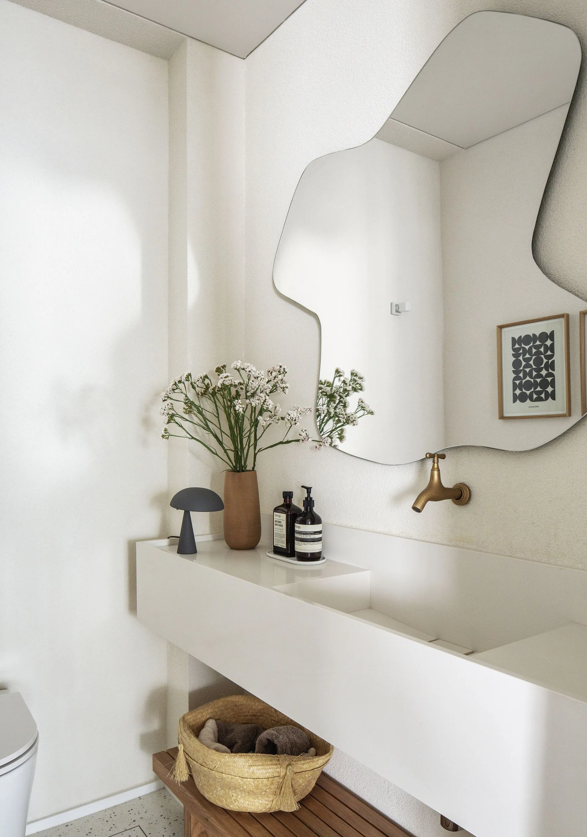 A wall-mounted white trough sink is paired with a brass faucet, an asymmetric mirror, and small decorative items on a textured plaster wall.