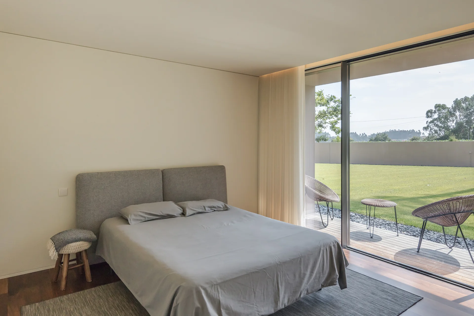 A minimalist bedroom features a grey bed next to a large sliding glass door that opens to a wooden patio and green lawn.