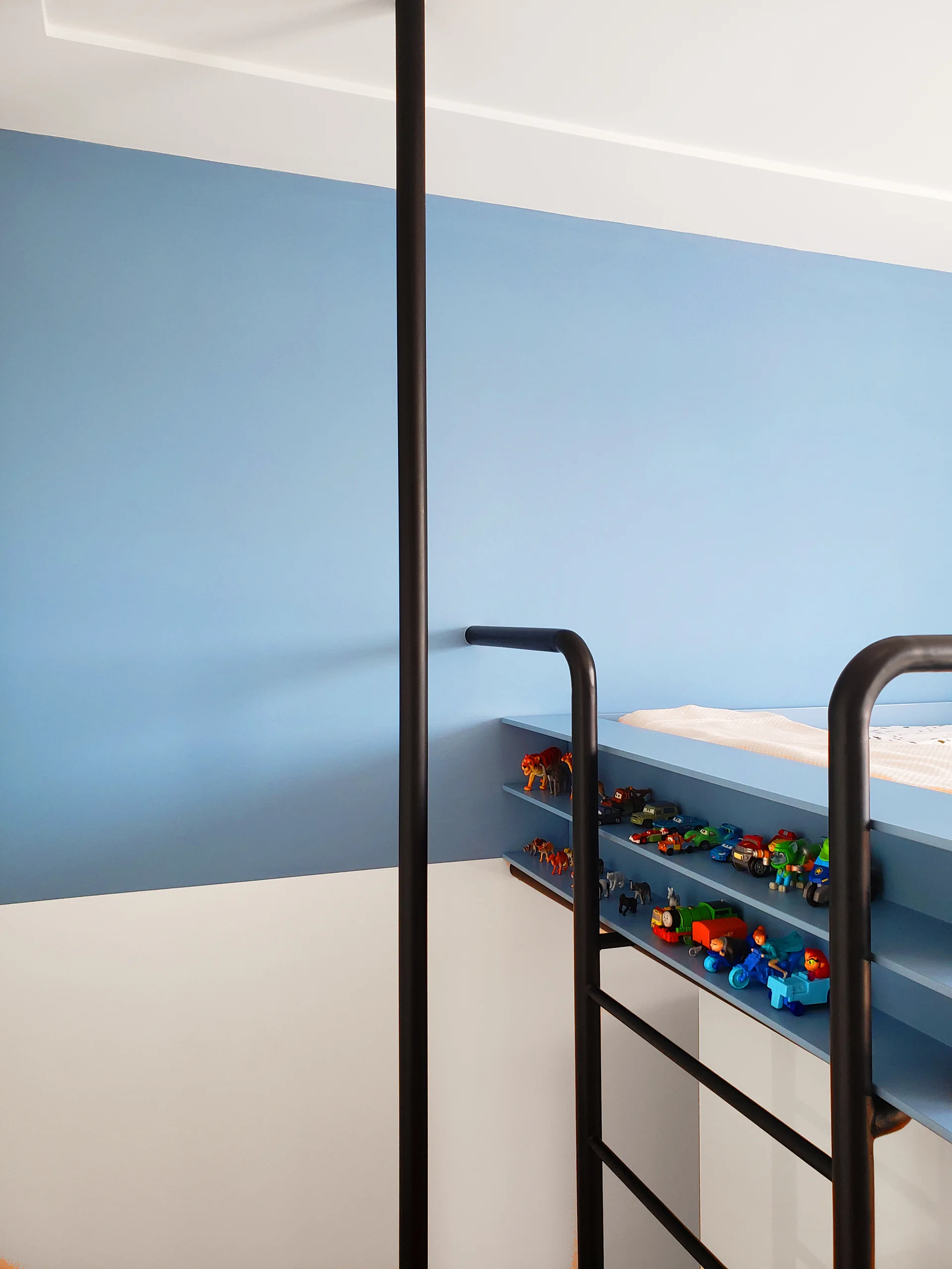 A blue bunk bed with a black metal ladder and built-in side shelves filled with small toys stands against a blue and white color-blocked wall.