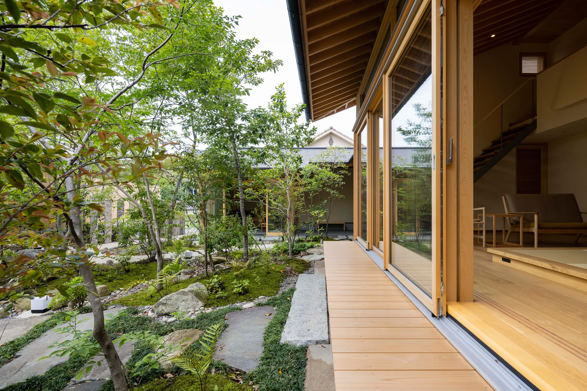 A light wood deck with large sliding glass doors opens to a lush green garden with stone pathways and moss.