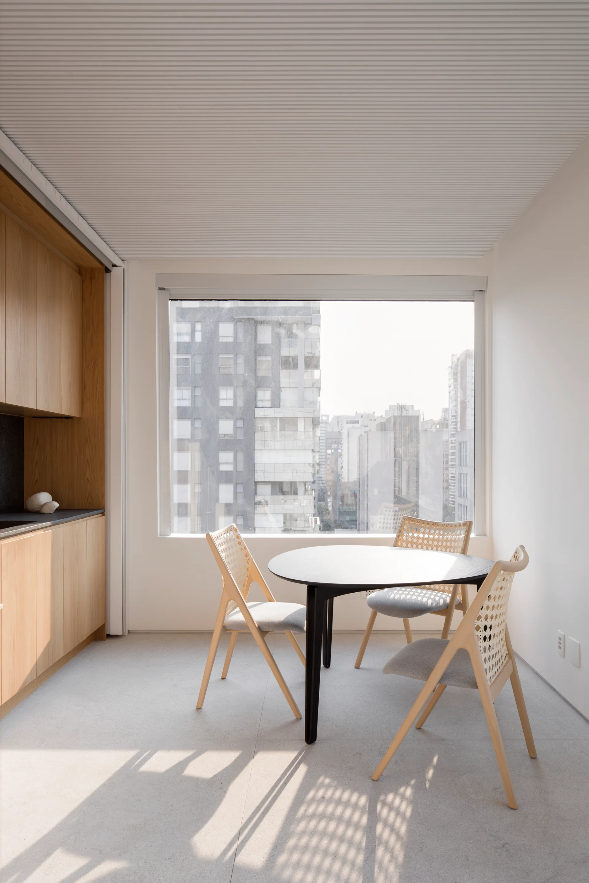 Three light wood chairs with caned backs surround a black round table next to a large window in a minimalist apartment.