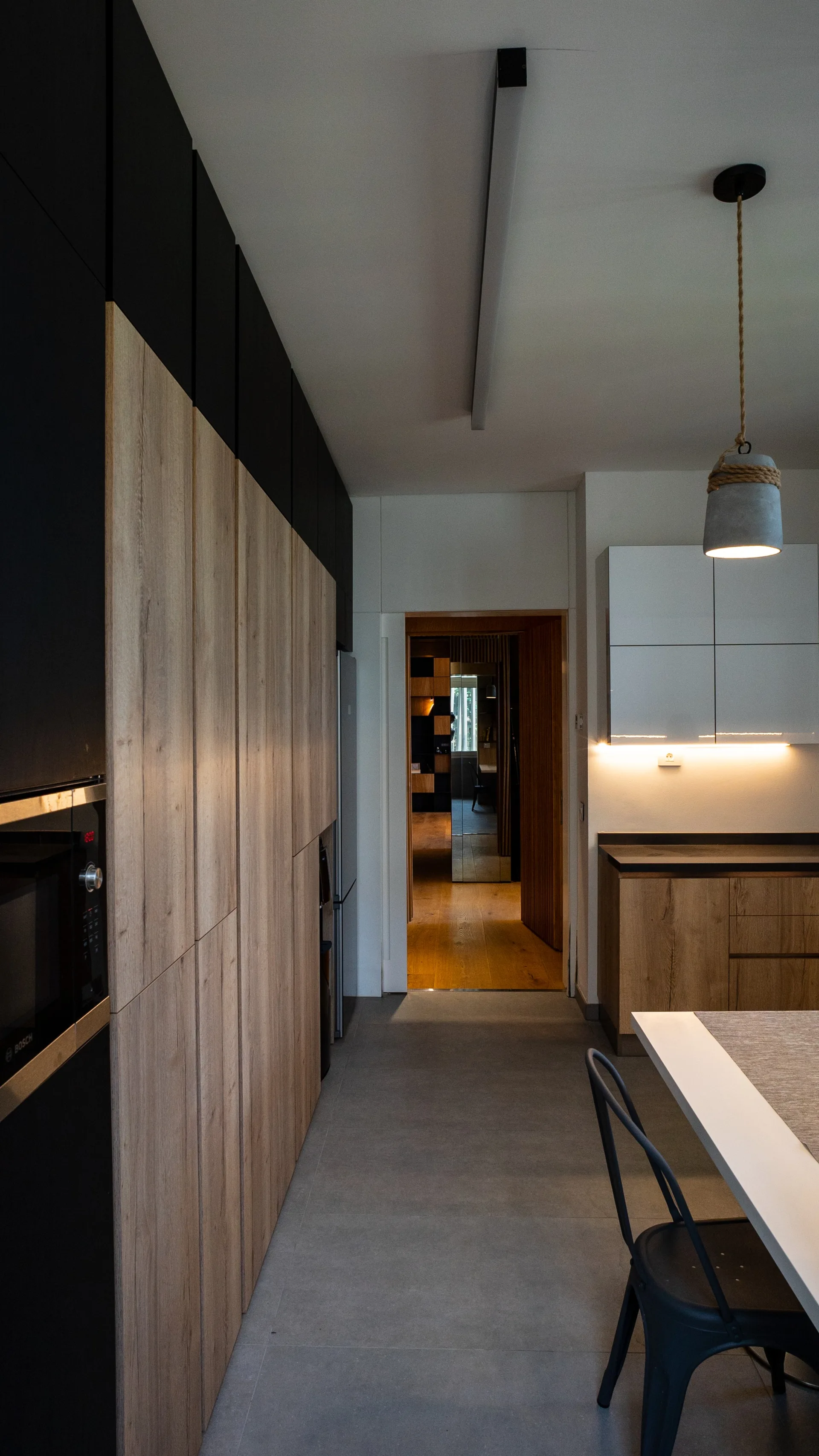 A modern kitchen features tall wood-grained and matte black cabinets, grey tile flooring, a dining table with a black chair, and a concrete pendant light.