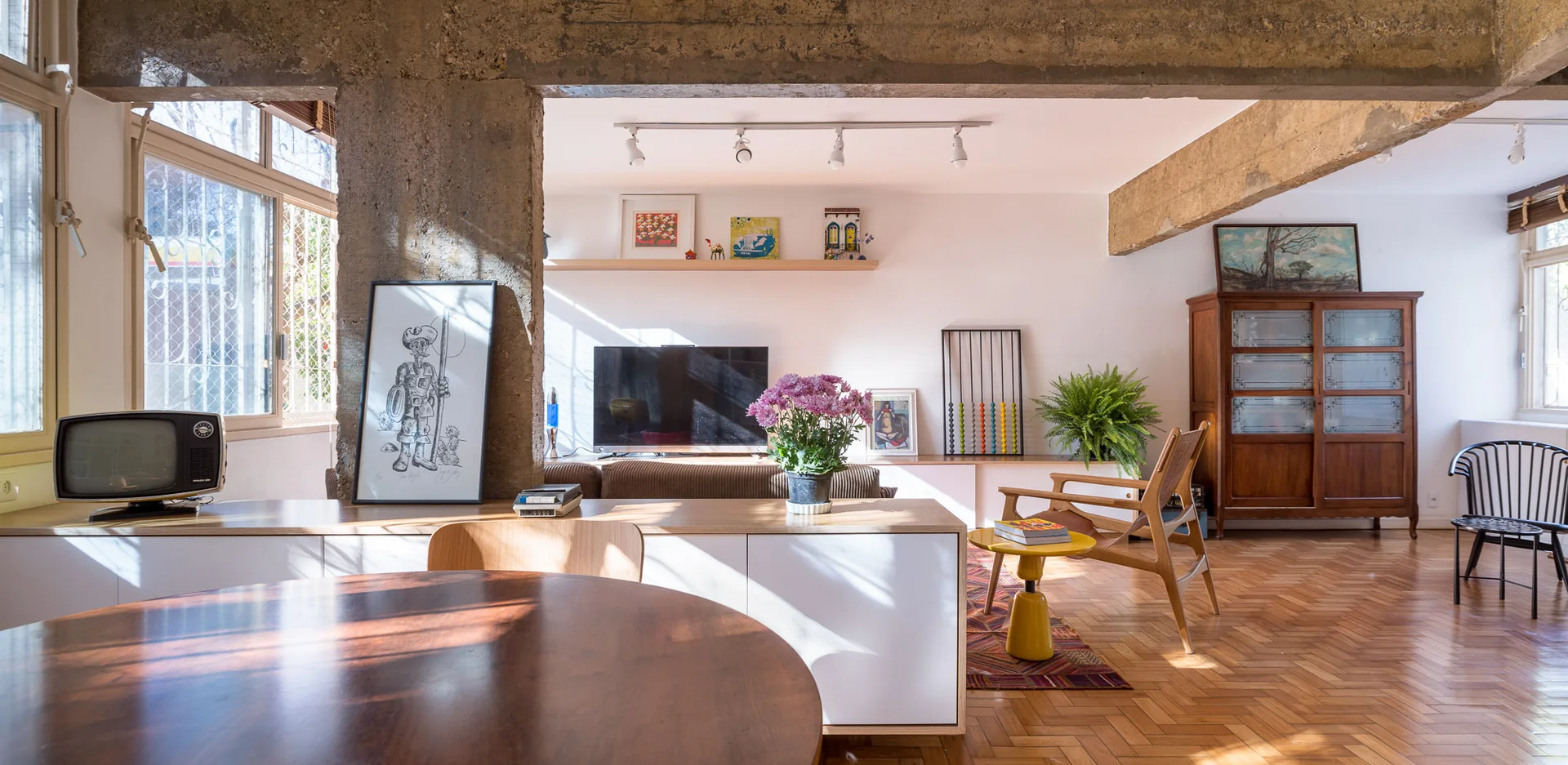 An open-plan living room features a herringbone wood parquet floor, raw concrete structural beams, low white cabinetry, and a round wooden dining table.