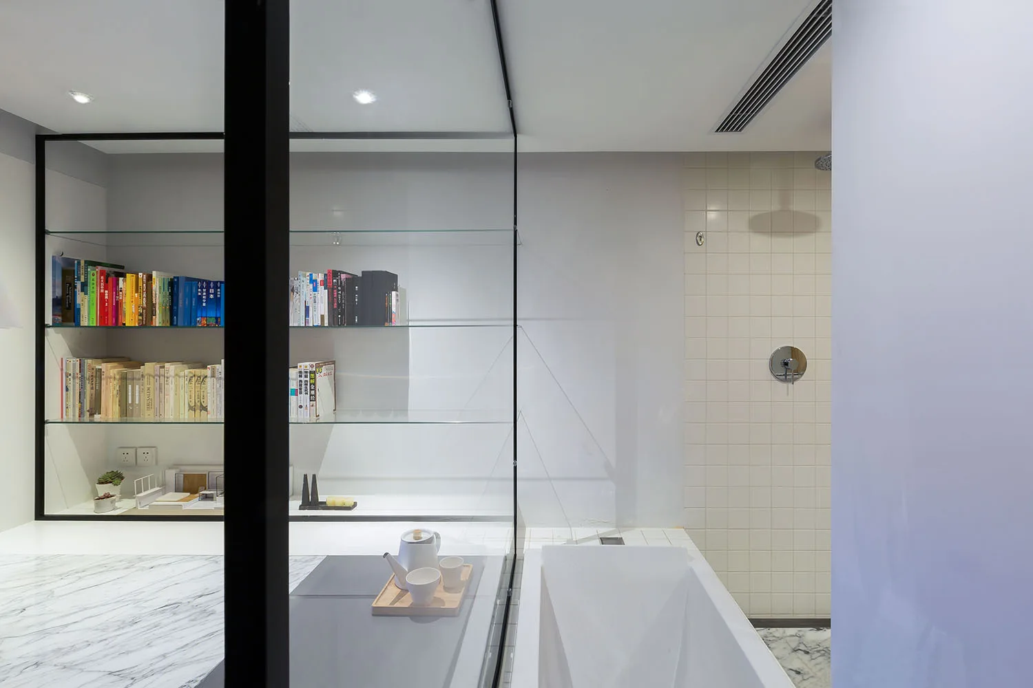 A black-framed glass shelving unit filled with books stands next to a white bathtub and tiled shower with marble flooring.