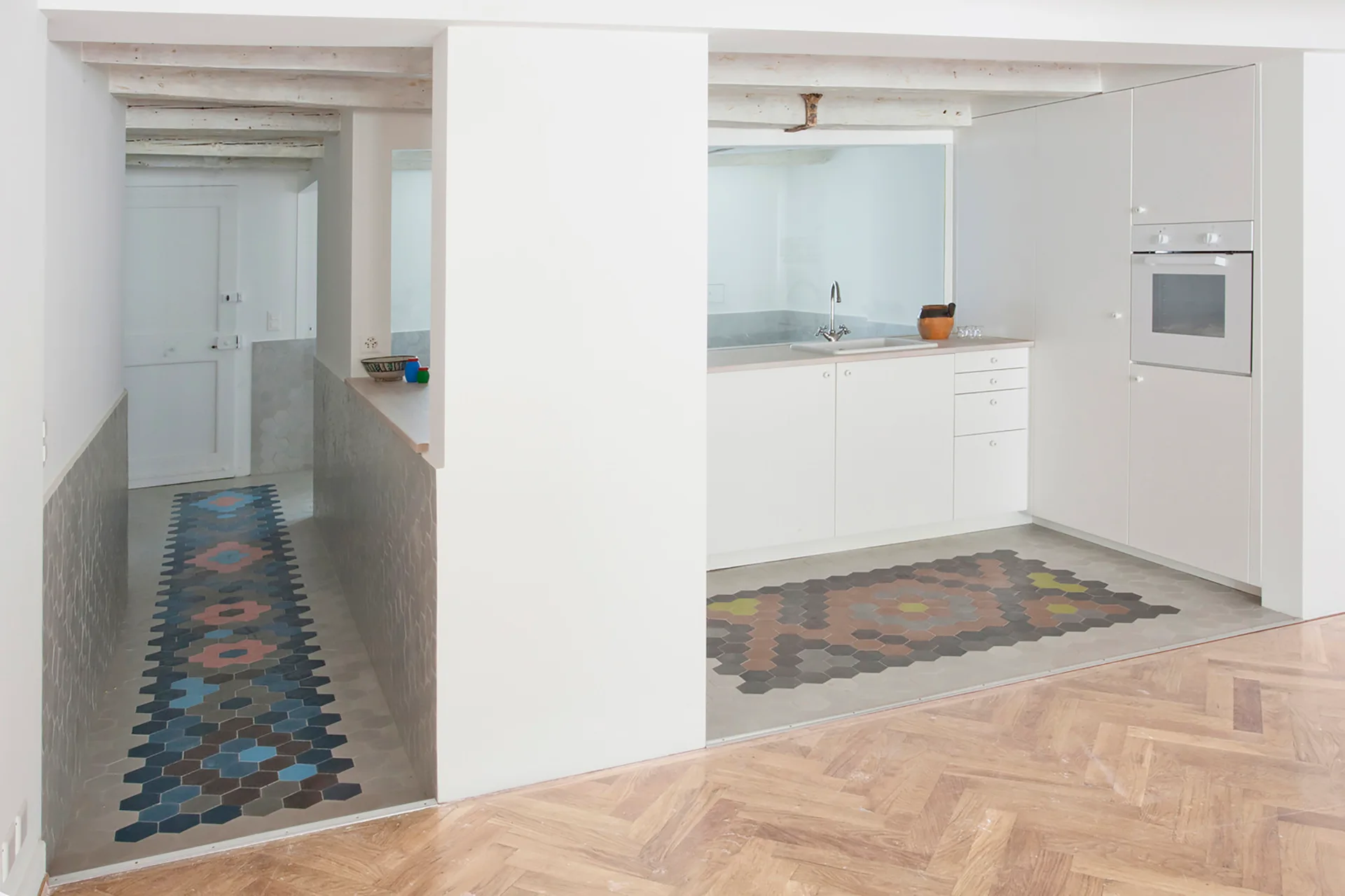 White minimalist kitchen and hallway featuring herringbone wood floors with colorful geometric hexagon tile inlays.