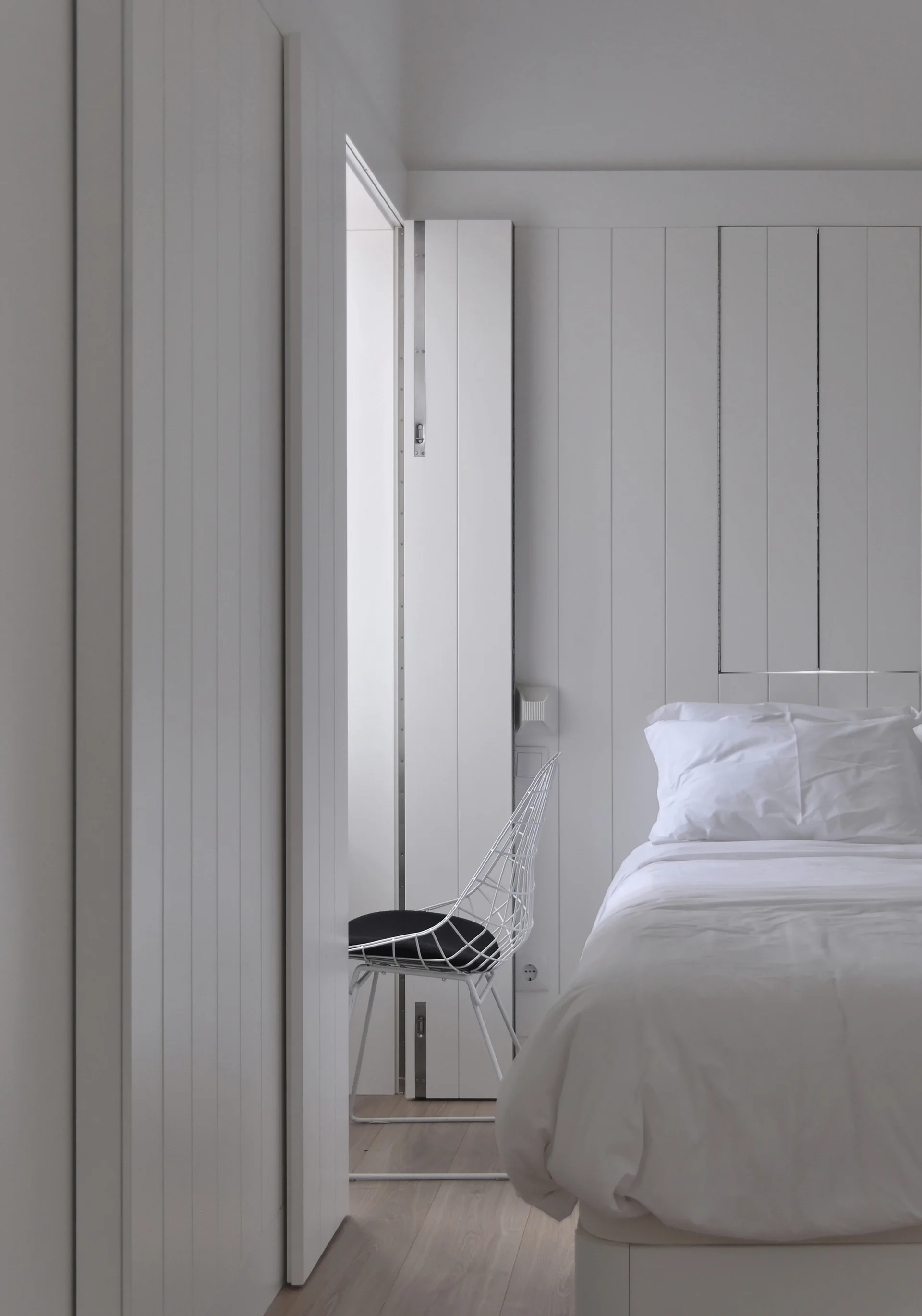 A minimalist bedroom featuring a bed with white linens next to vertical wall paneling and a wireframe chair in the doorway.