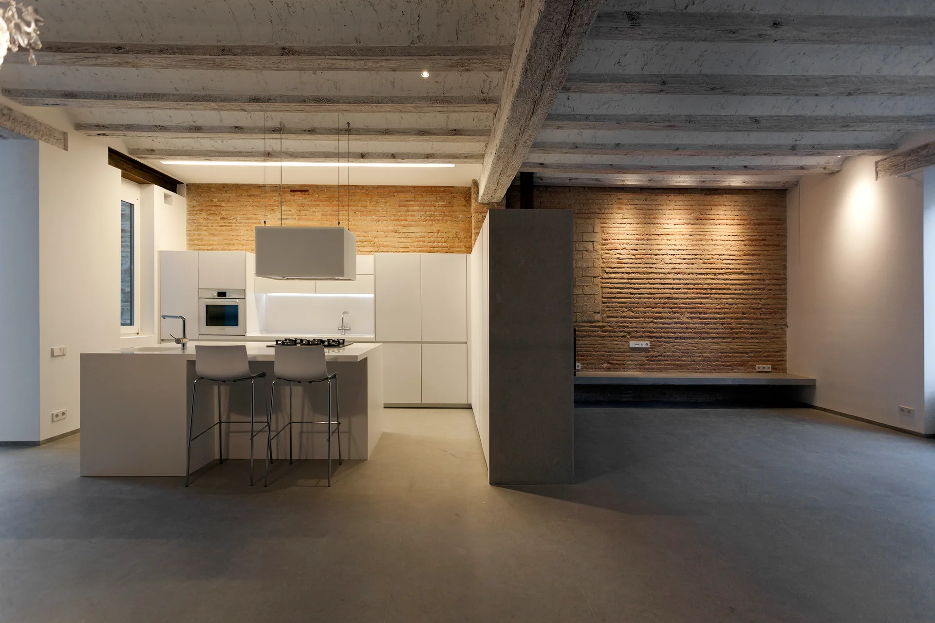 A minimalist white kitchen island and cabinets stand against an exposed brick wall under a rustic wooden beamed ceiling with concrete floors.
