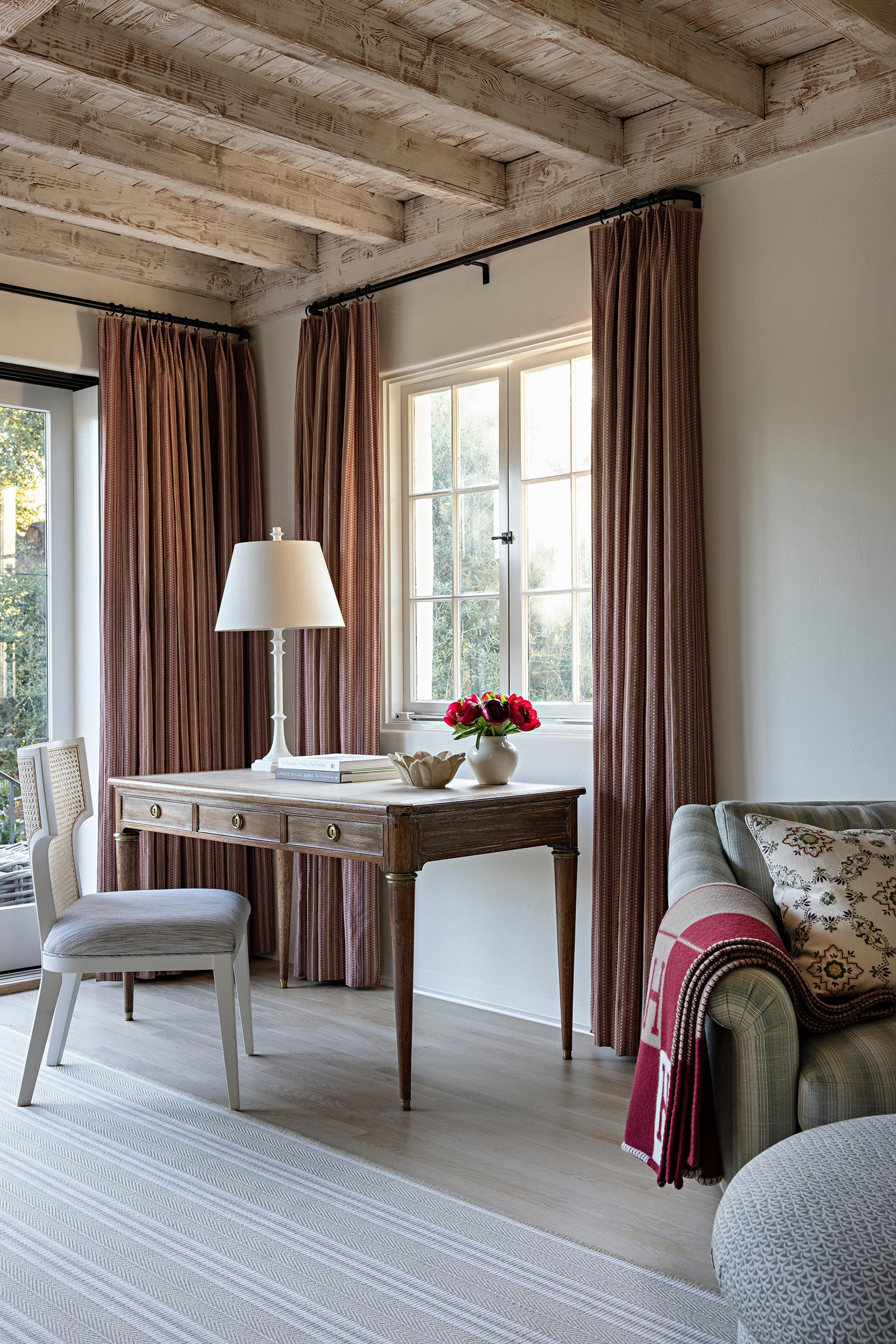 A wooden writing desk with a white lamp and chair stands in front of a window framed by terracotta pinstripe curtains beneath an exposed wood ceiling.