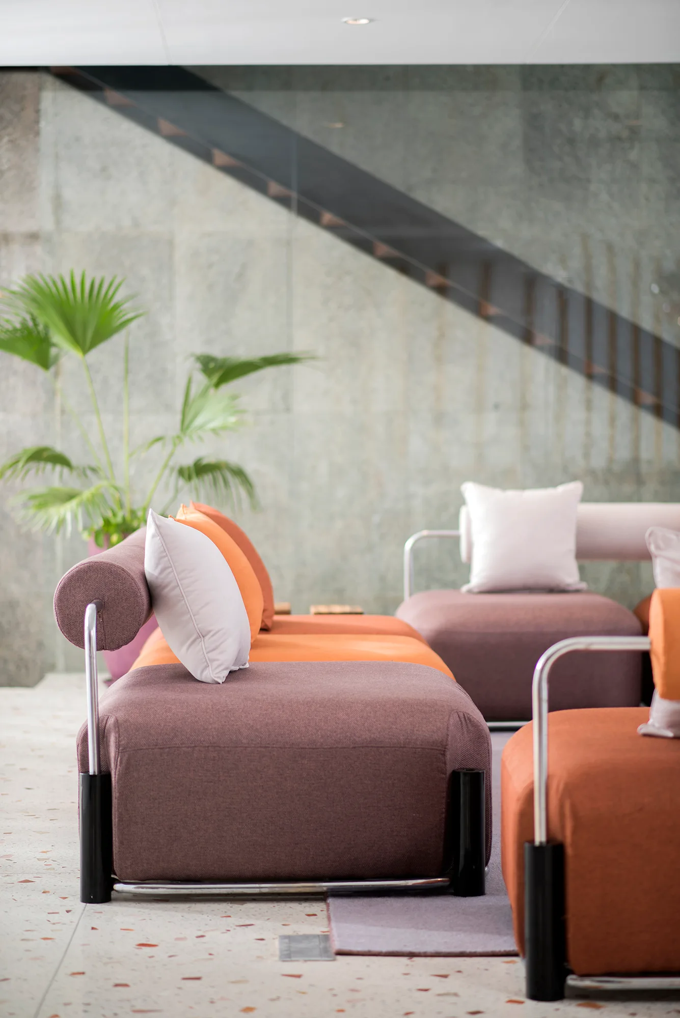 A modern lounge area with low-slung modular sofa cushions in terracotta and plum, set against a grey stone wall and terrazzo flooring.