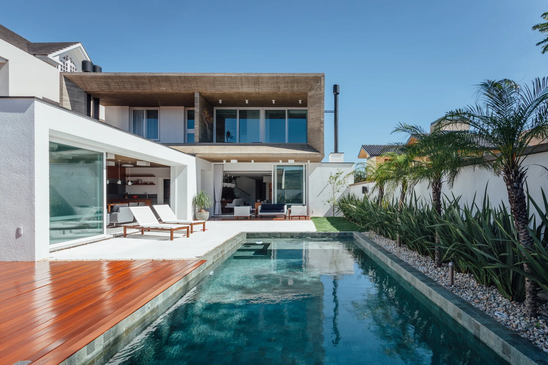 A rectangular backyard swimming pool sits next to a modern two-story home with a wood deck, white outdoor lounge chairs, and an upper concrete canopy.