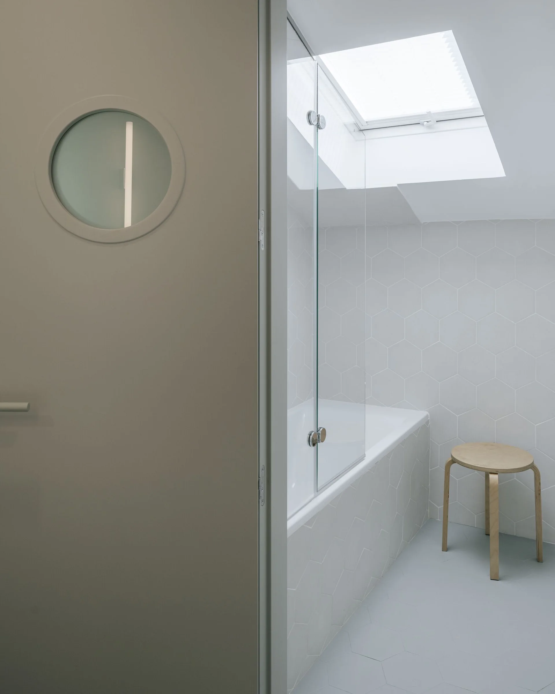 A minimalist bathroom featuring a grey door with a round porthole window, a bathtub surrounded by grey hexagon tiles, and a skylight overhead.