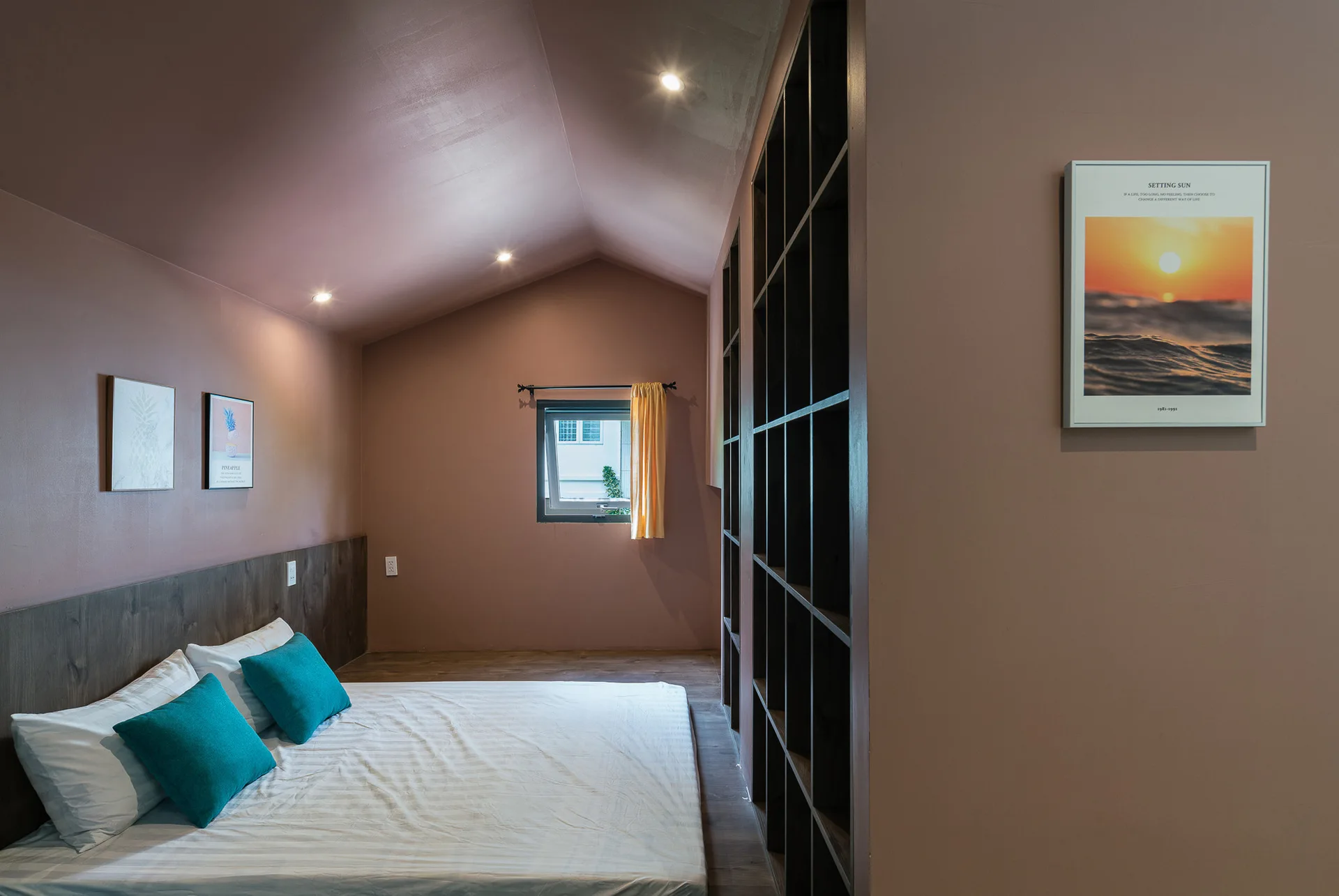 A mauve attic bedroom featuring a low platform bed with teal pillows, a tall dark wood shelving unit, and a sloped ceiling.